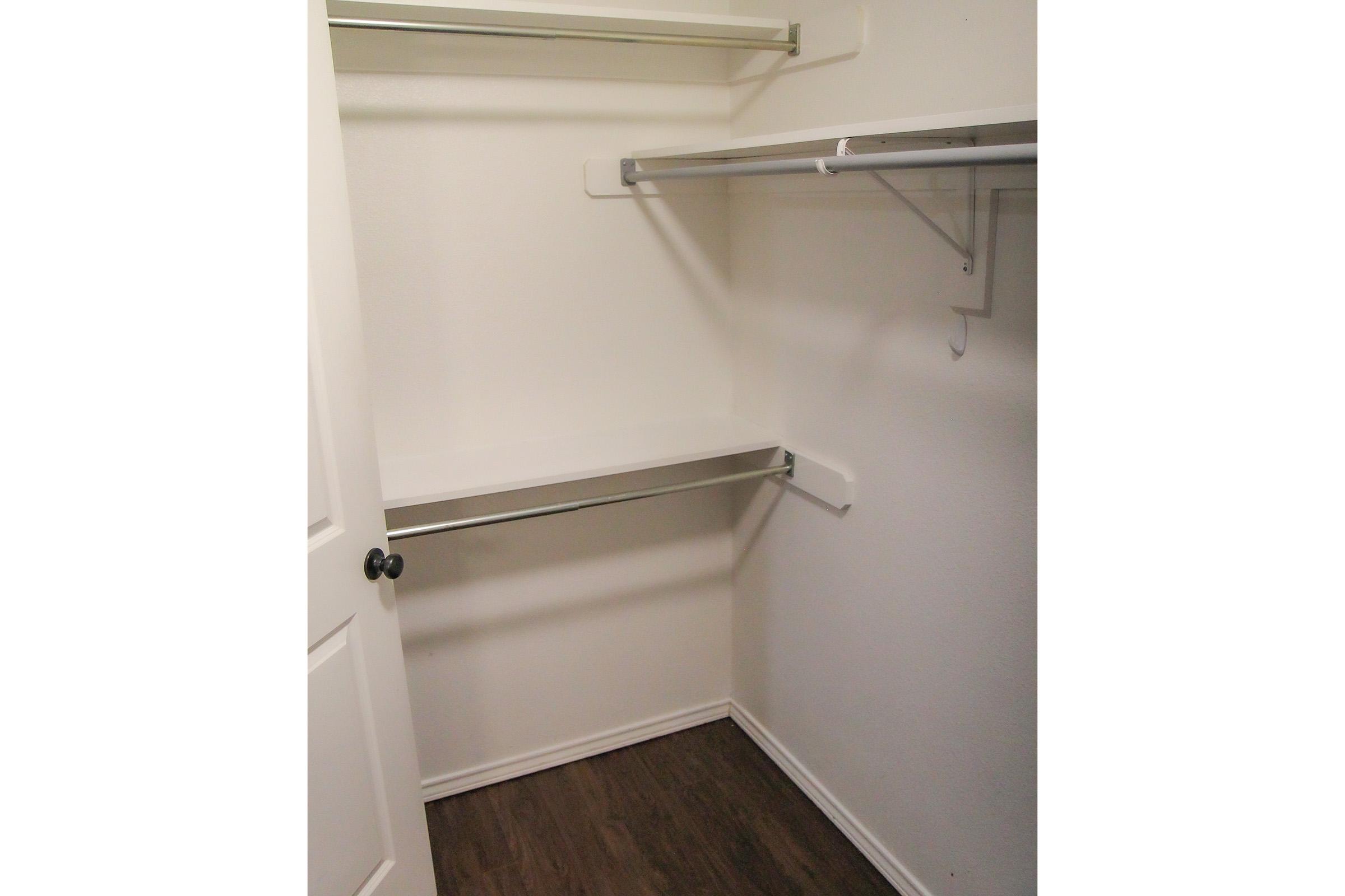 Empty closet with white walls, a wooden floor, and two horizontal shelving units mounted on the sides. There is a shelf above the floor and a hanging rod for clothes. The door is closed, and the overall space appears organized and clean.