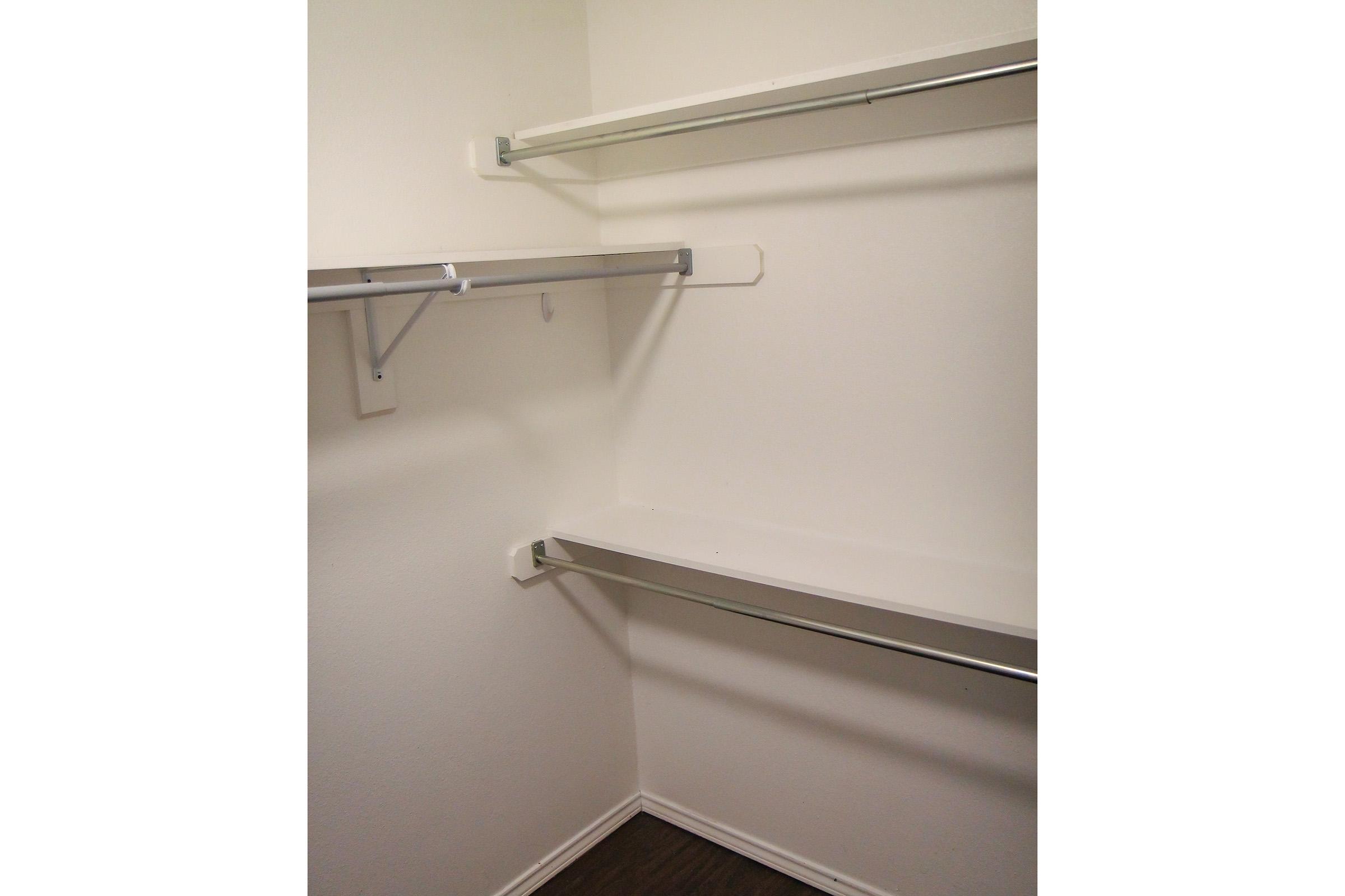 Empty closet with white walls and two metal shelves mounted on the sides. There is a single shelf along the back wall, all surfaces clean and unadorned, with dark wooden flooring visible below.