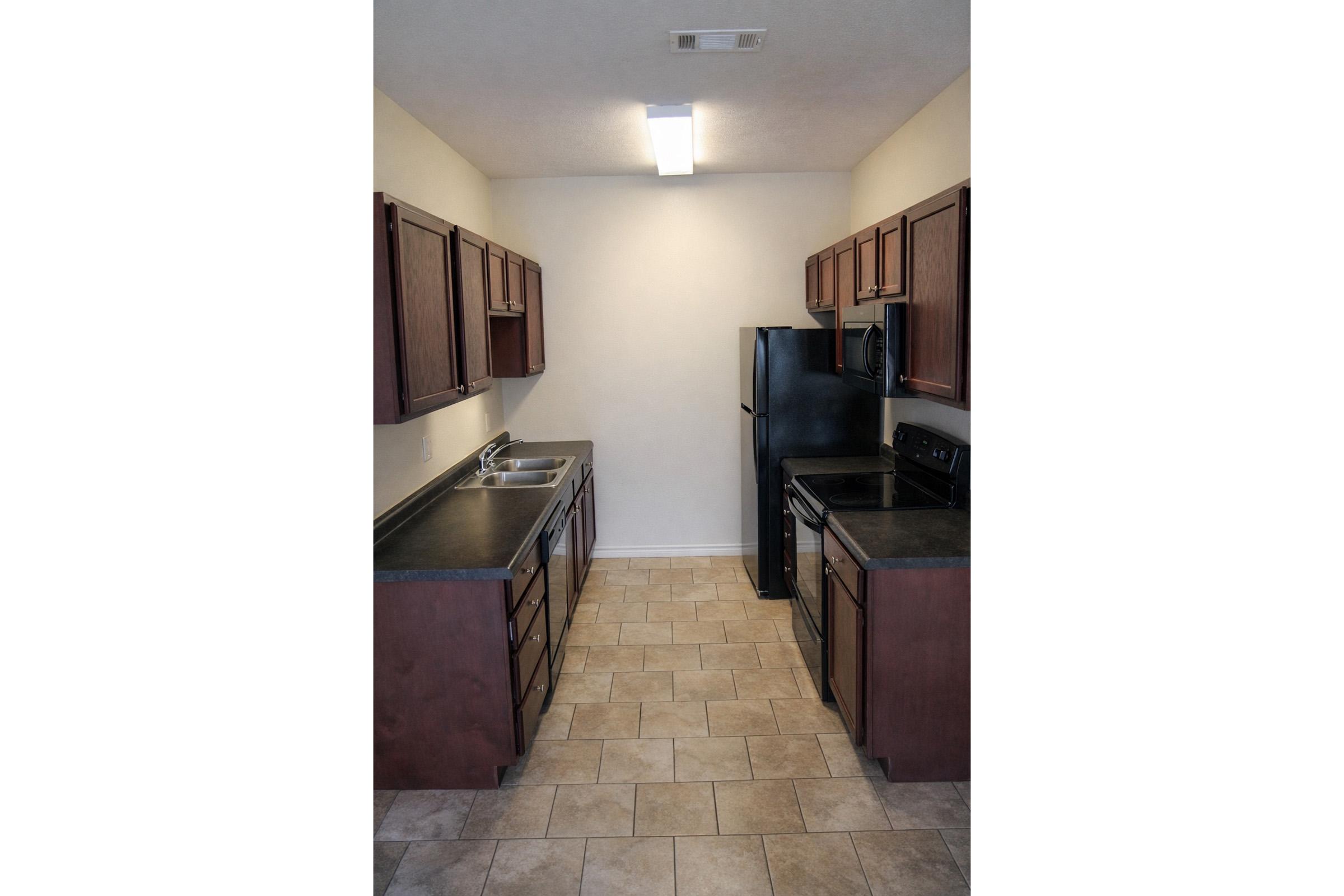 A modern kitchen featuring dark wood cabinets, black appliances including a refrigerator and oven, a sink with a countertop, and tiled flooring. The walls are painted a light color, and there is overhead lighting providing brightness to the space.