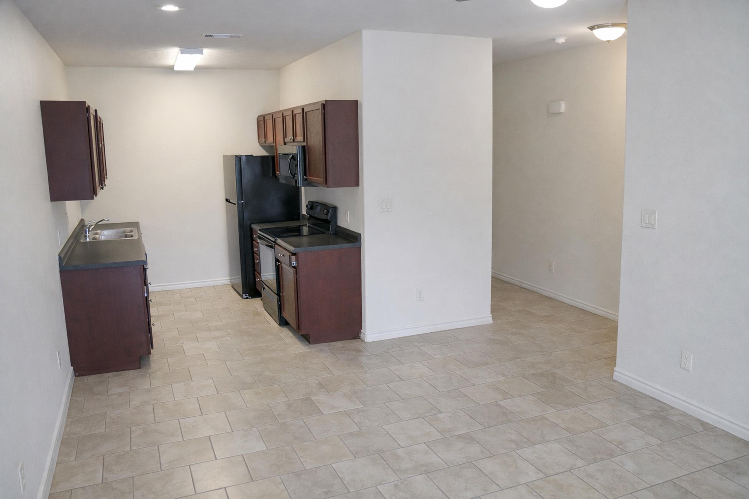 A spacious kitchen area featuring dark wooden cabinets, a black refrigerator, and a stove. The open layout leads to a well-lit living space with tiled flooring, white walls, and minimal decor, providing a modern and clean aesthetic.