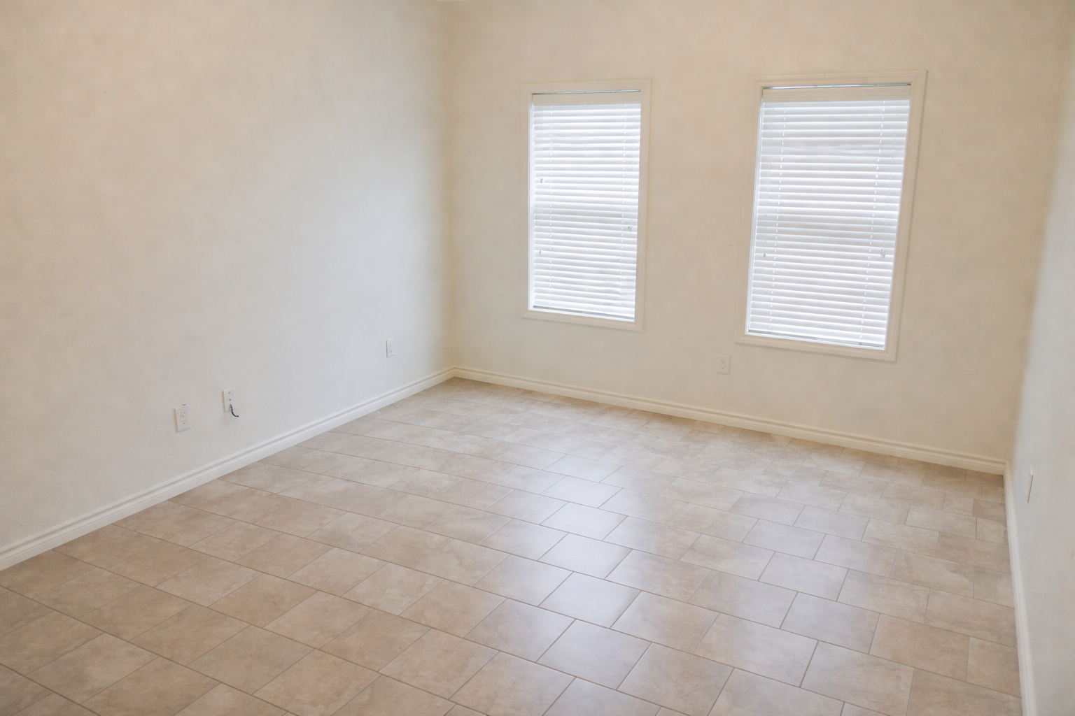 A spacious, empty room featuring light-colored walls and two windows with blinds. The floor is covered in beige tiles, providing a clean and minimalist aesthetic. The overall ambiance is bright and airy, ideal for various uses such as a living space or office.