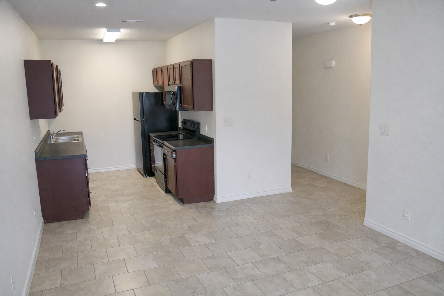 A modern, open-concept kitchen and living area featuring light-colored walls, tiled flooring, dark wood cabinets, and stainless steel appliances, including a refrigerator and stove. The space is well-lit with natural light and has an uncluttered, clean aesthetic.