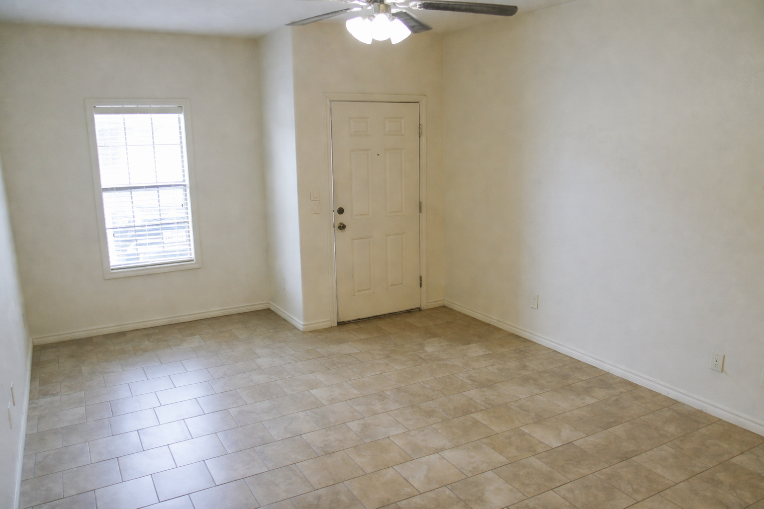 An empty room with light beige walls and tiled flooring. There is a window with blinds on the left side, and a closed door with a doorknob on the right. A ceiling fan is visible above, and the space appears well-lit, creating a simple and open atmosphere.