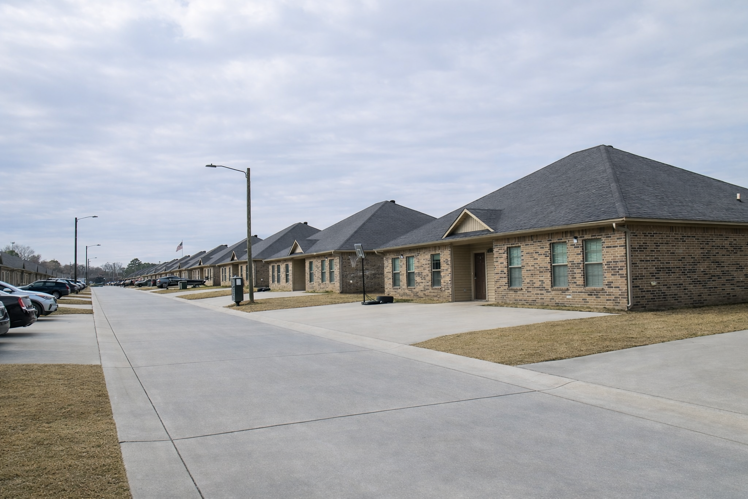 Row of single-story brick houses with sloped roofs, lined along a wide concrete street. The scene features a cloudy sky and well-kept lawns. Parked cars are visible along the street, adding to the residential atmosphere.
