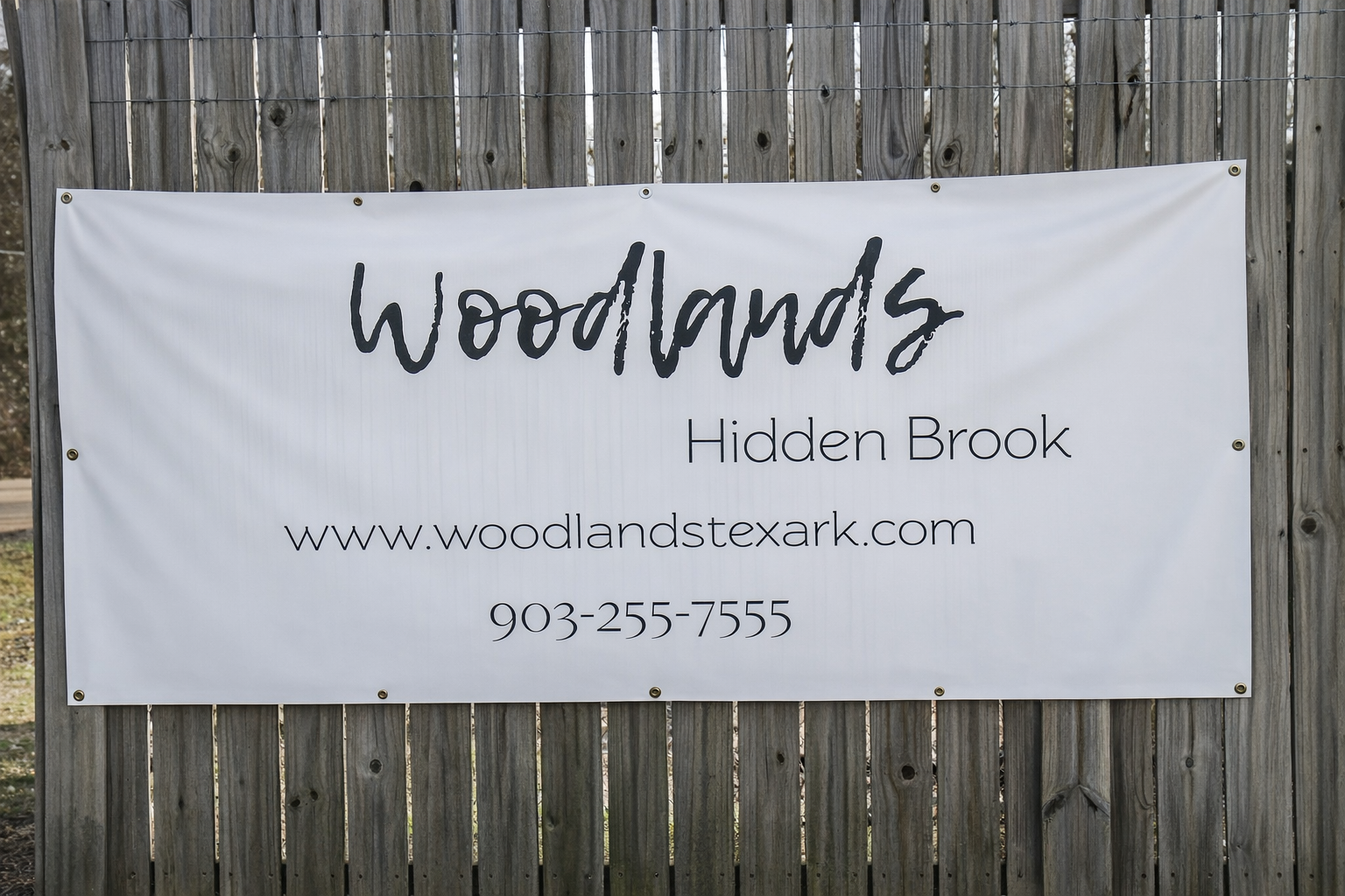 A large banner on a wooden fence displaying the text "Woodlands Hidden Brook" along with a website URL "www.woodlandstexark.com" and a phone number "903-255-7555." The banner has a clean design with a modern font.