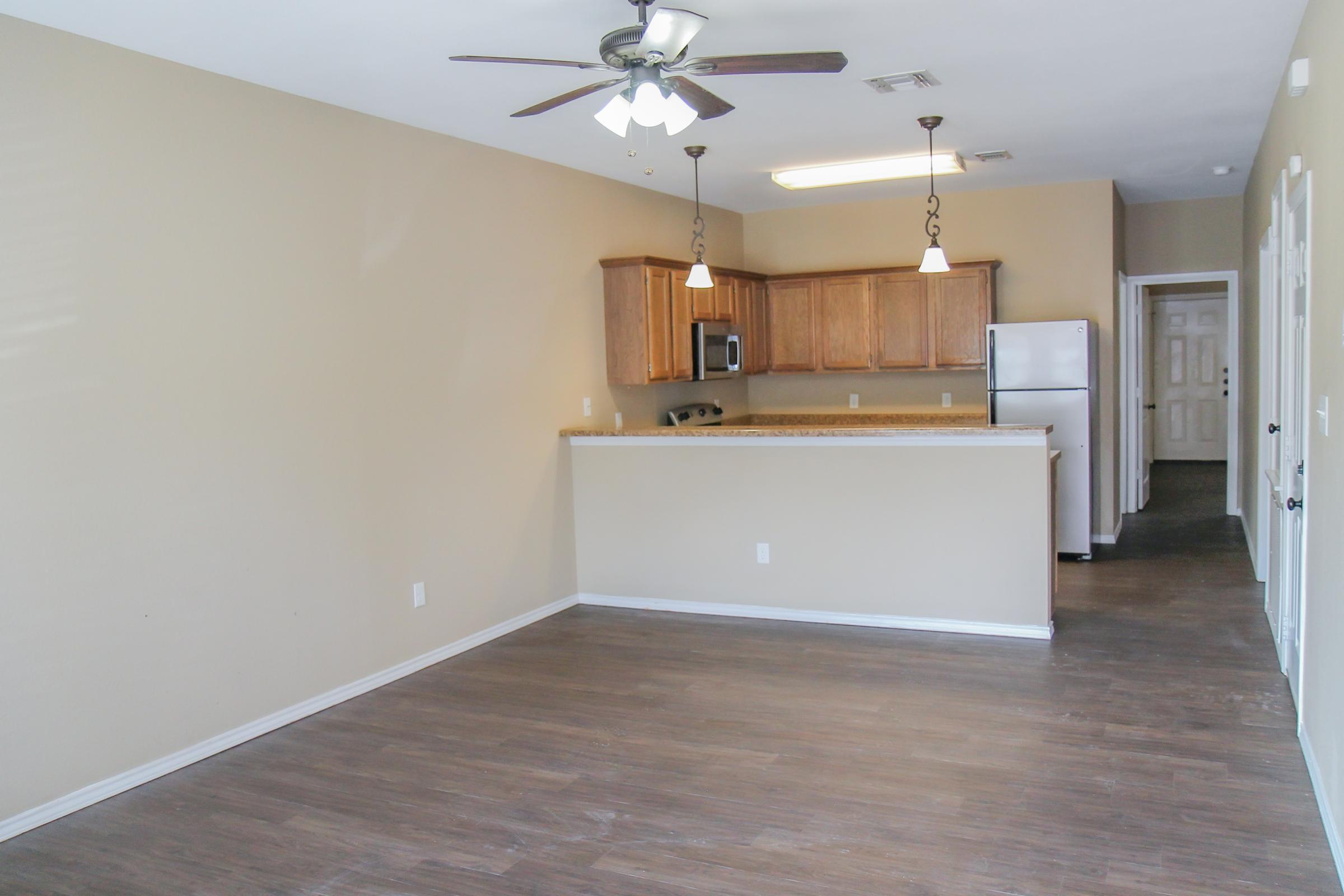Interior view of a modern, vacant apartment featuring a spacious living area with laminate flooring. An open kitchen with wooden cabinets, a countertop, and stainless steel appliances is visible. The walls are painted in a light beige color, with a ceiling fan and decorative light fixtures. Doors lead to other areas of the apartment.