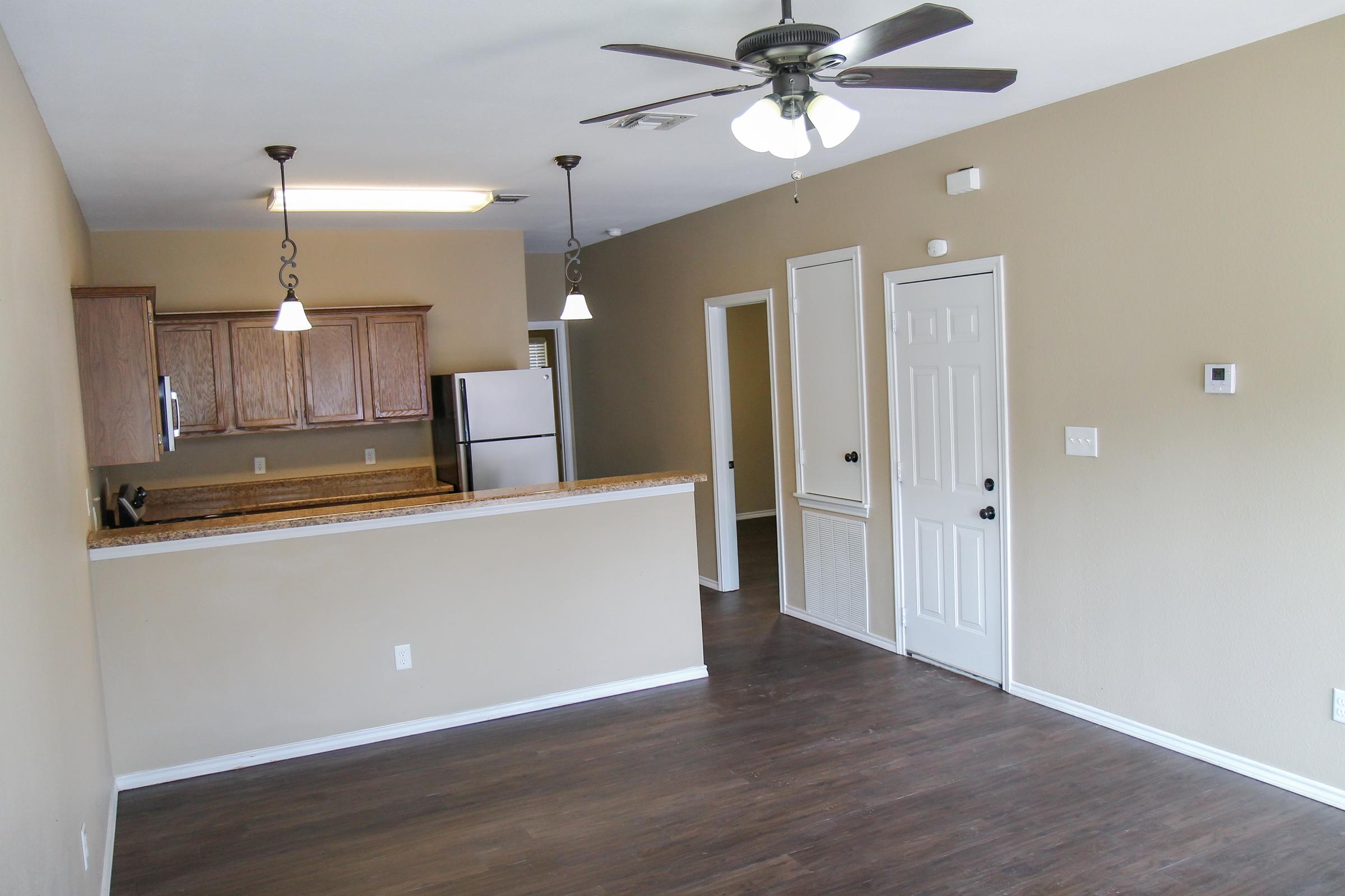 A spacious, light-filled living area with wood laminate flooring, featuring an open layout that connects to a kitchen with wooden cabinets and stainless steel appliances. There are two ceiling fans, a door leading outside, and a doorway leading to another room, all set against a neutral beige wall color.