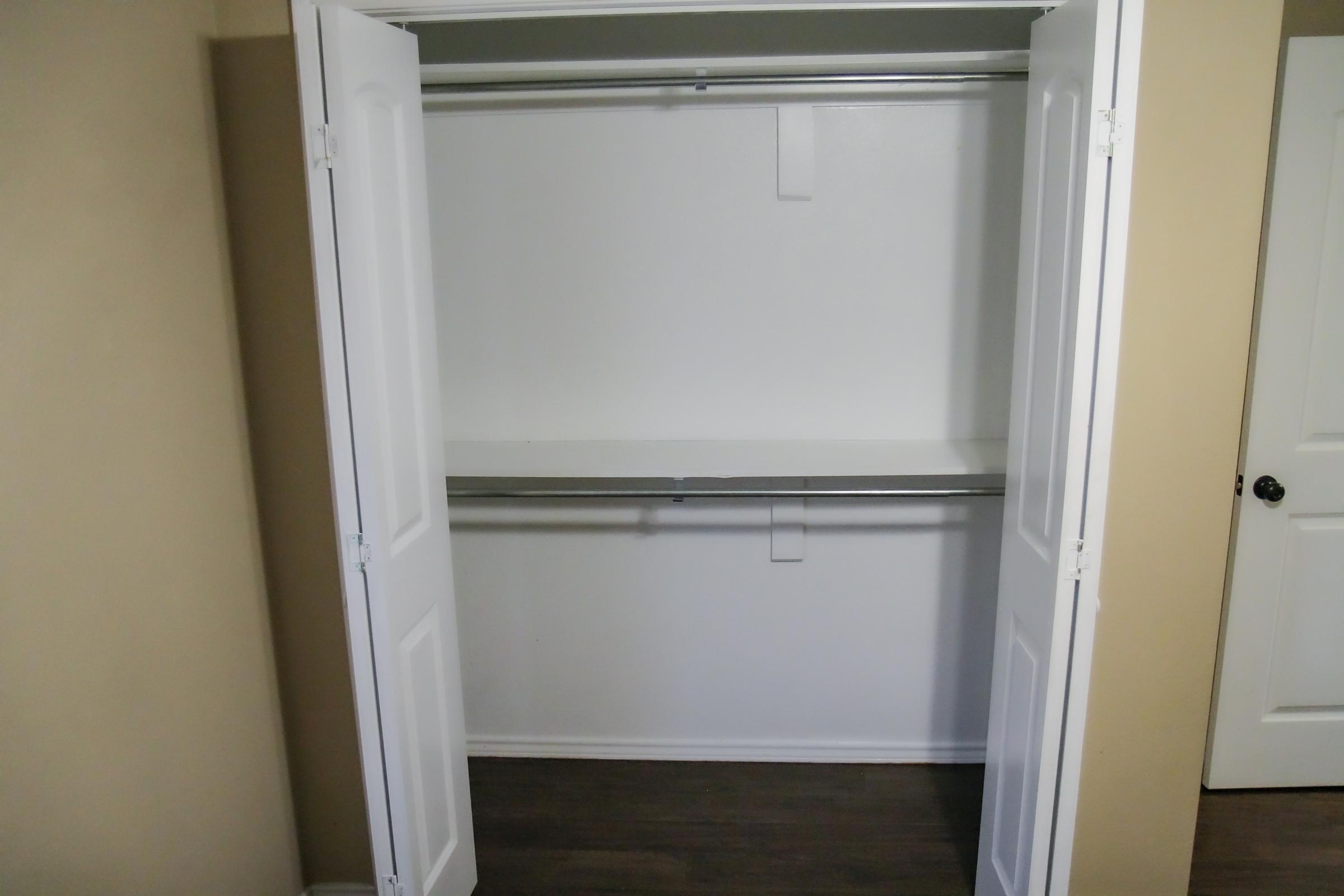 A clean, empty closet with double white doors slightly open, revealing a simple interior with a single shelf and a clothing rod. The walls are painted white, and the floor features dark wood. A door is visible to the right of the closet.