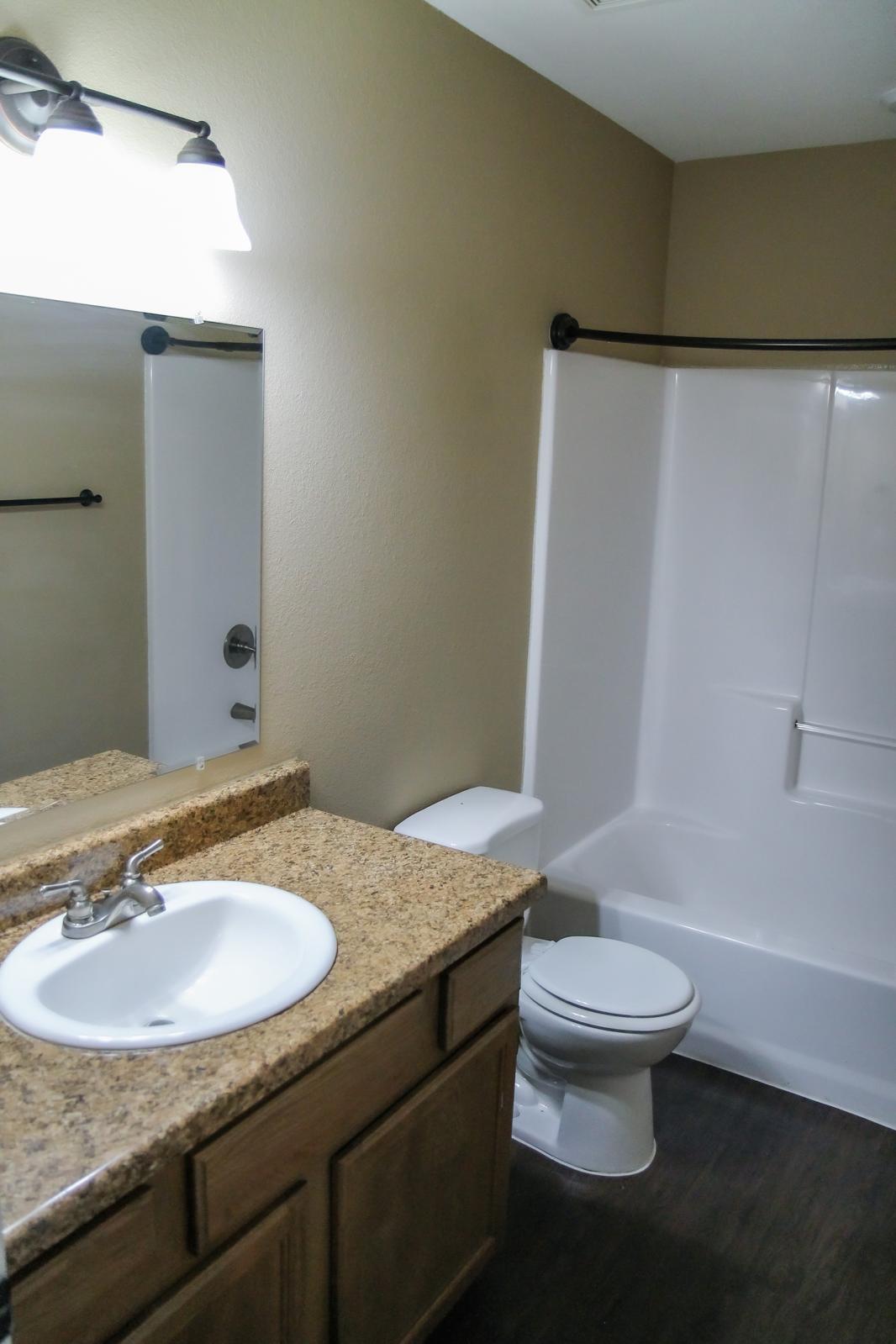 A clean and well-lit bathroom featuring a granite countertop sink with a mirror above, a toilet, and a shower area with a white tub and glass door. The walls are painted in a warm beige color, and there is a towel bar on the wall. The flooring is dark, contrasting with the lighter fixtures.