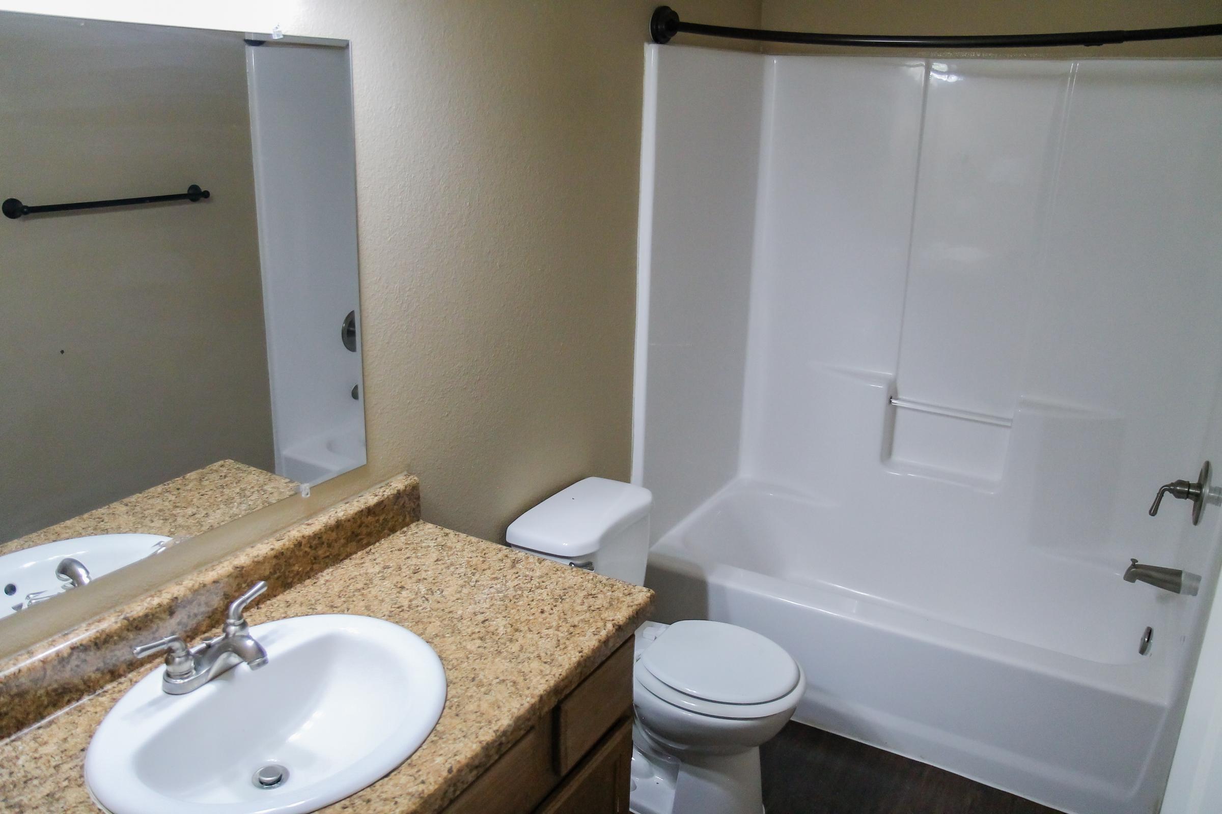 A clean bathroom featuring a white bathtub and shower, a sink with a granite countertop, and a toilet. The walls are painted a light color, and there is a mirror above the sink. A towel rack is visible on the wall, and the flooring is dark. Natural light comes from a window beside the mirror.
