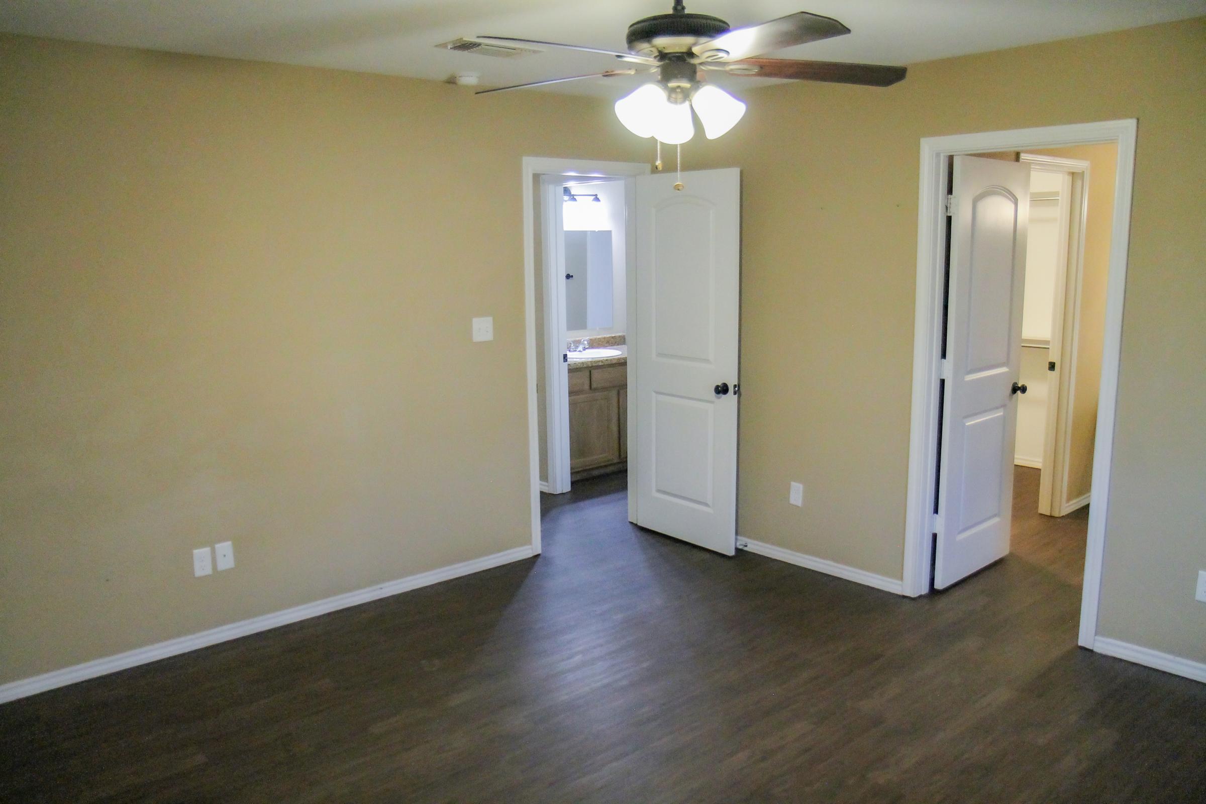 A simple interior of a room featuring light brown walls and laminate flooring. There are two doors leading to adjacent spaces, one likely to a bathroom with a sink visible, and the other to another room. A ceiling fan hangs from the ceiling, providing a bright and open atmosphere.
