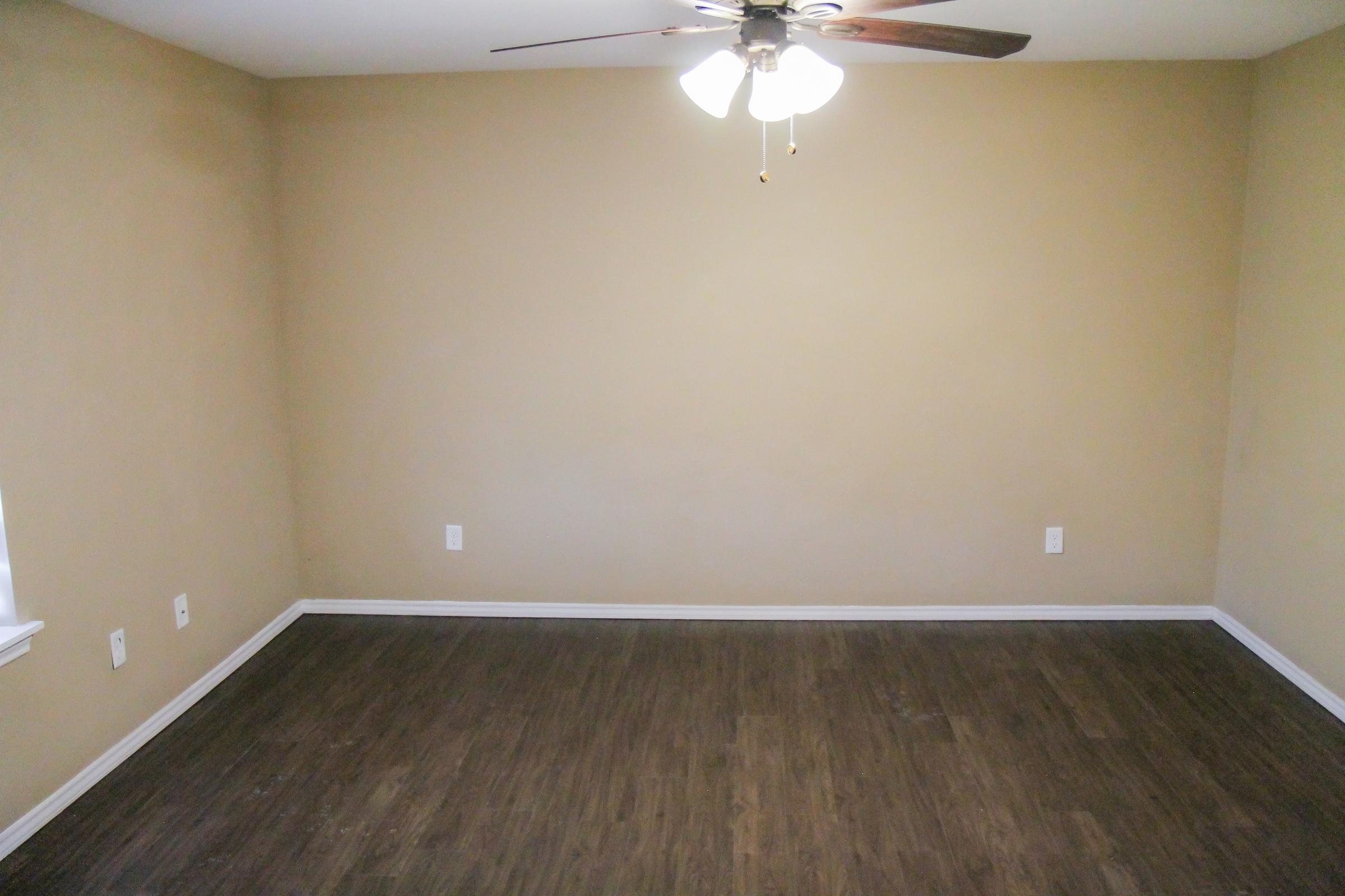 Empty room with light brown walls, a ceiling fan with five blades, and dark wooden flooring. There are no furniture or decorations present. Two electrical outlets are visible on the walls, and a window is located on one side, allowing natural light to enter the space.