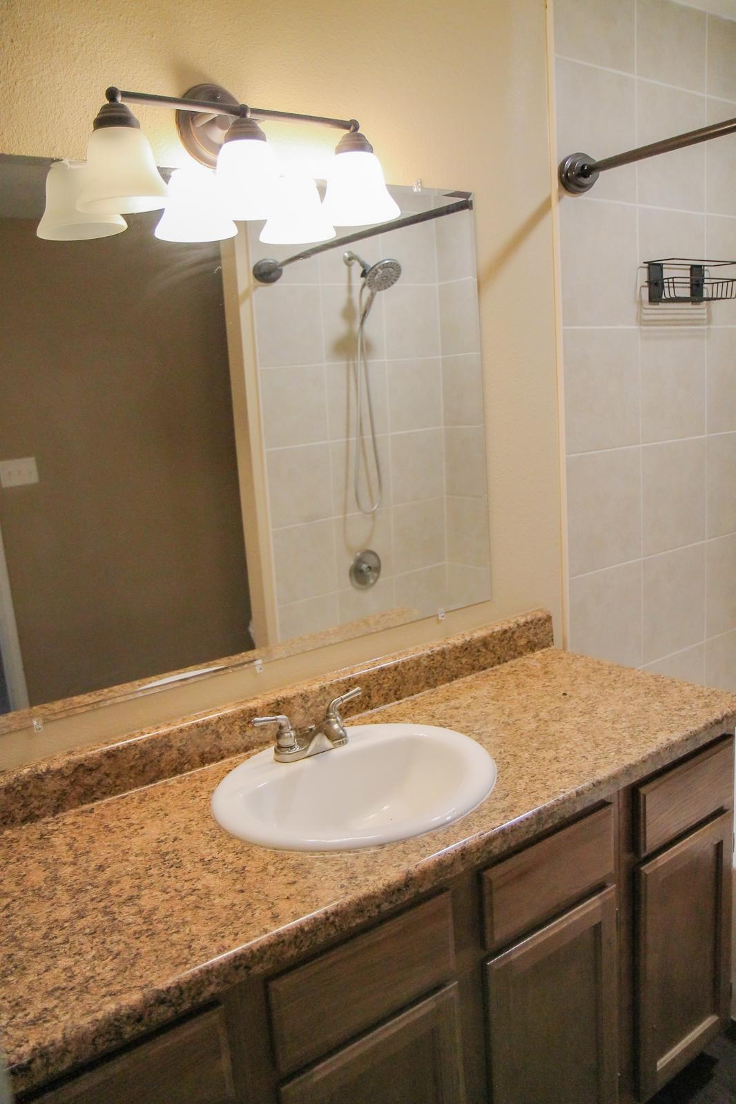 A bathroom vanity with a granite countertop, an oval sink, and a modern faucet. Above the sink, there is a mirror with a light fixture featuring three glass shades. A shower area is visible in the background, along with tiled walls and a showerhead. The overall color scheme is neutral and clean.