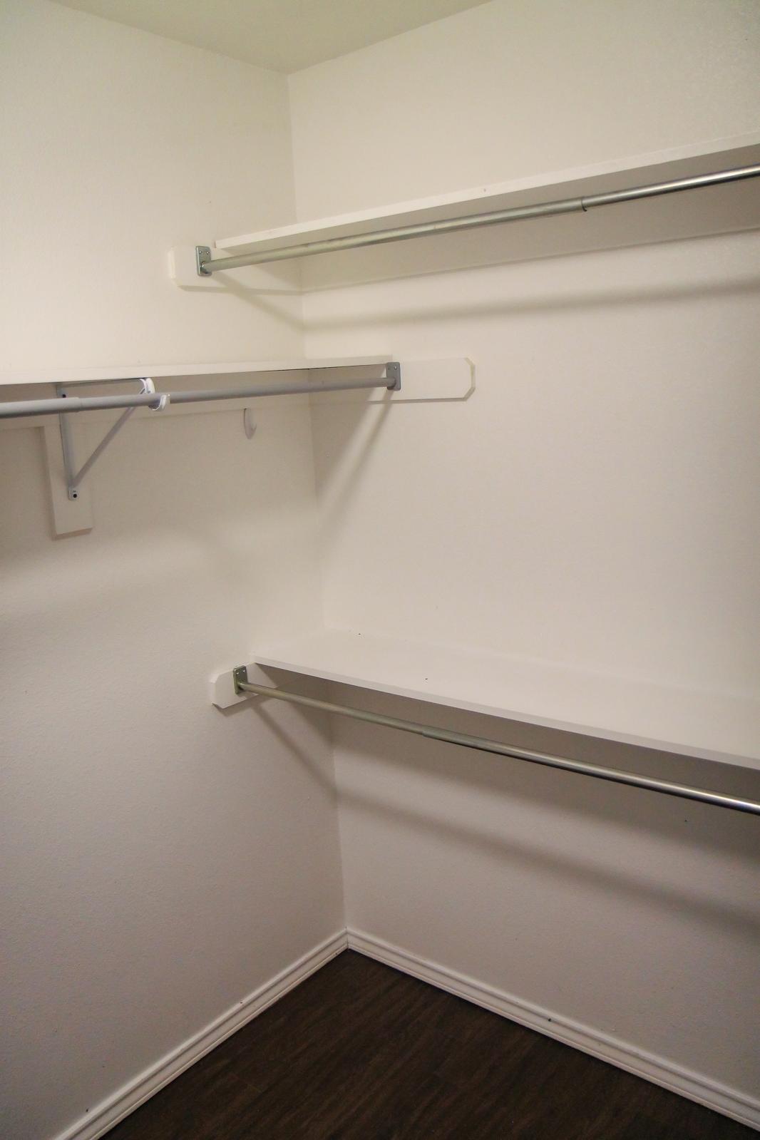 An empty closet featuring two metal shelves and a hook on one wall, with plain white walls and a dark wood floor. The minimalistic design showcases ample space for storage or organization.