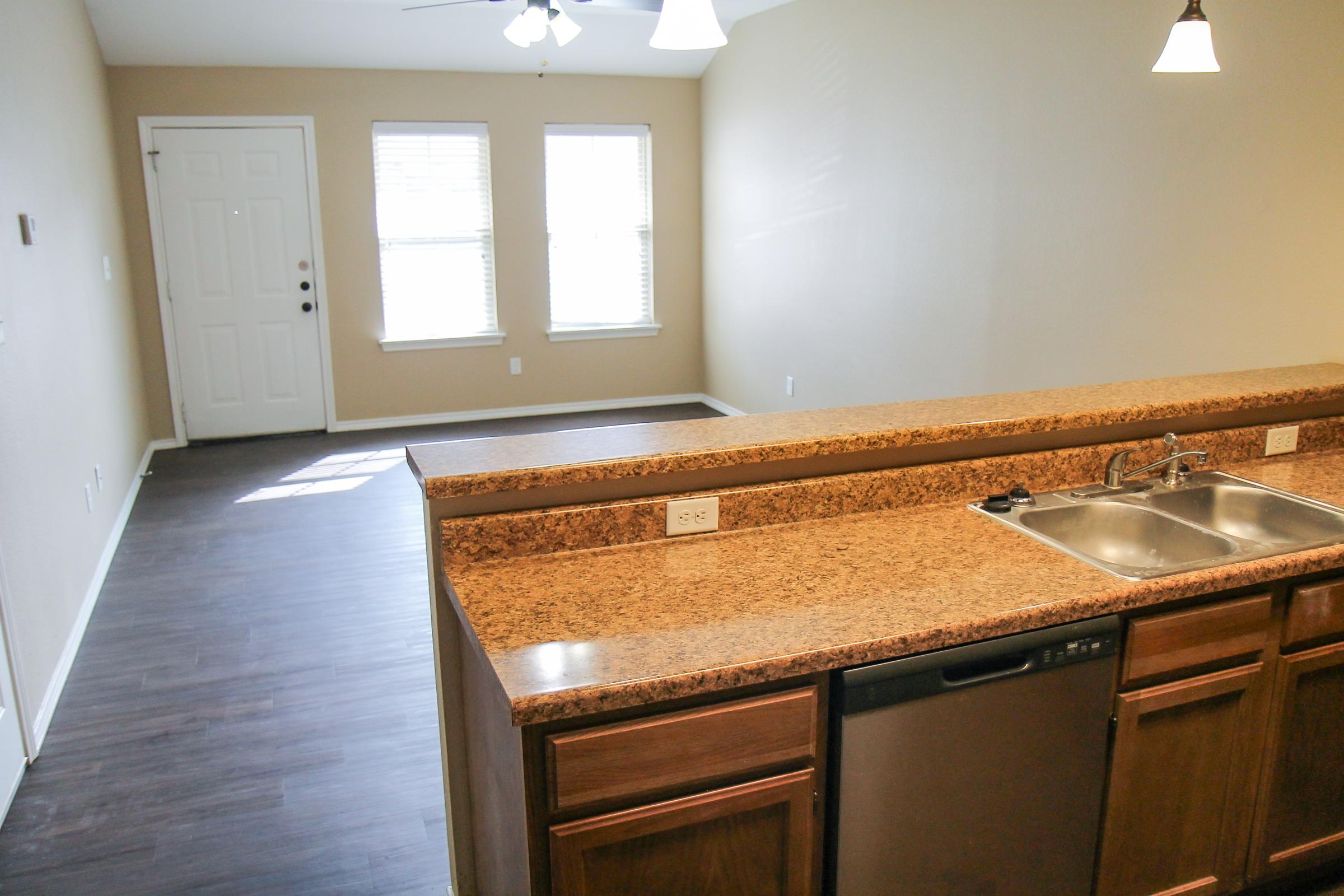 A modern kitchen with a granite countertop, a double sink, and a dishwasher. The background features a spacious living area with two windows allowing natural light, and a front door visible. The flooring is dark wood, and the walls are painted a soft beige color. Pendant lights hang above the countertop.