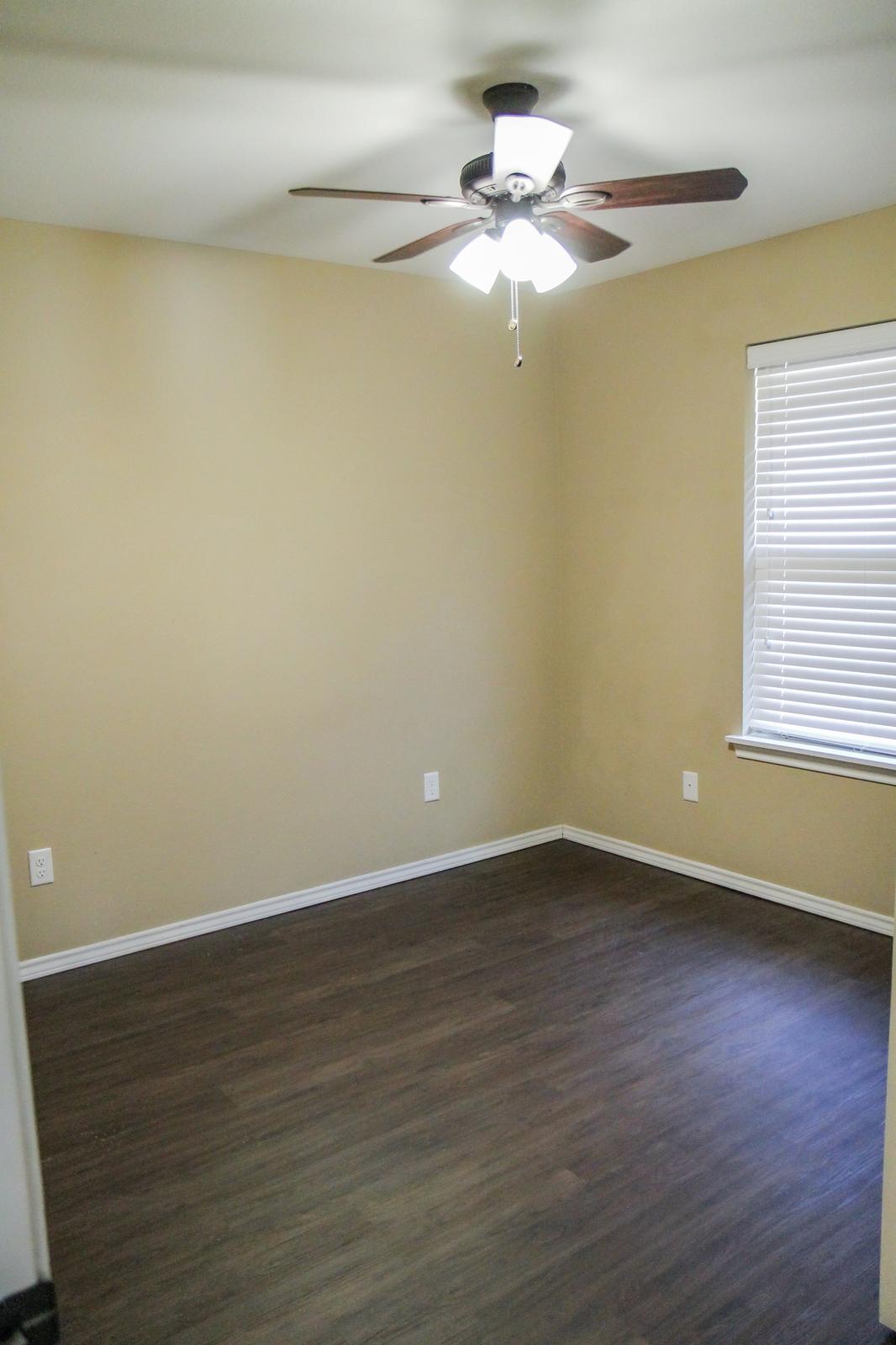 A well-lit empty room with beige walls and a ceiling fan. The floor is wooden, and there is a window with blinds on the right side, allowing natural light to enter. The room has no furniture or decorations, creating a spacious and uncluttered atmosphere.