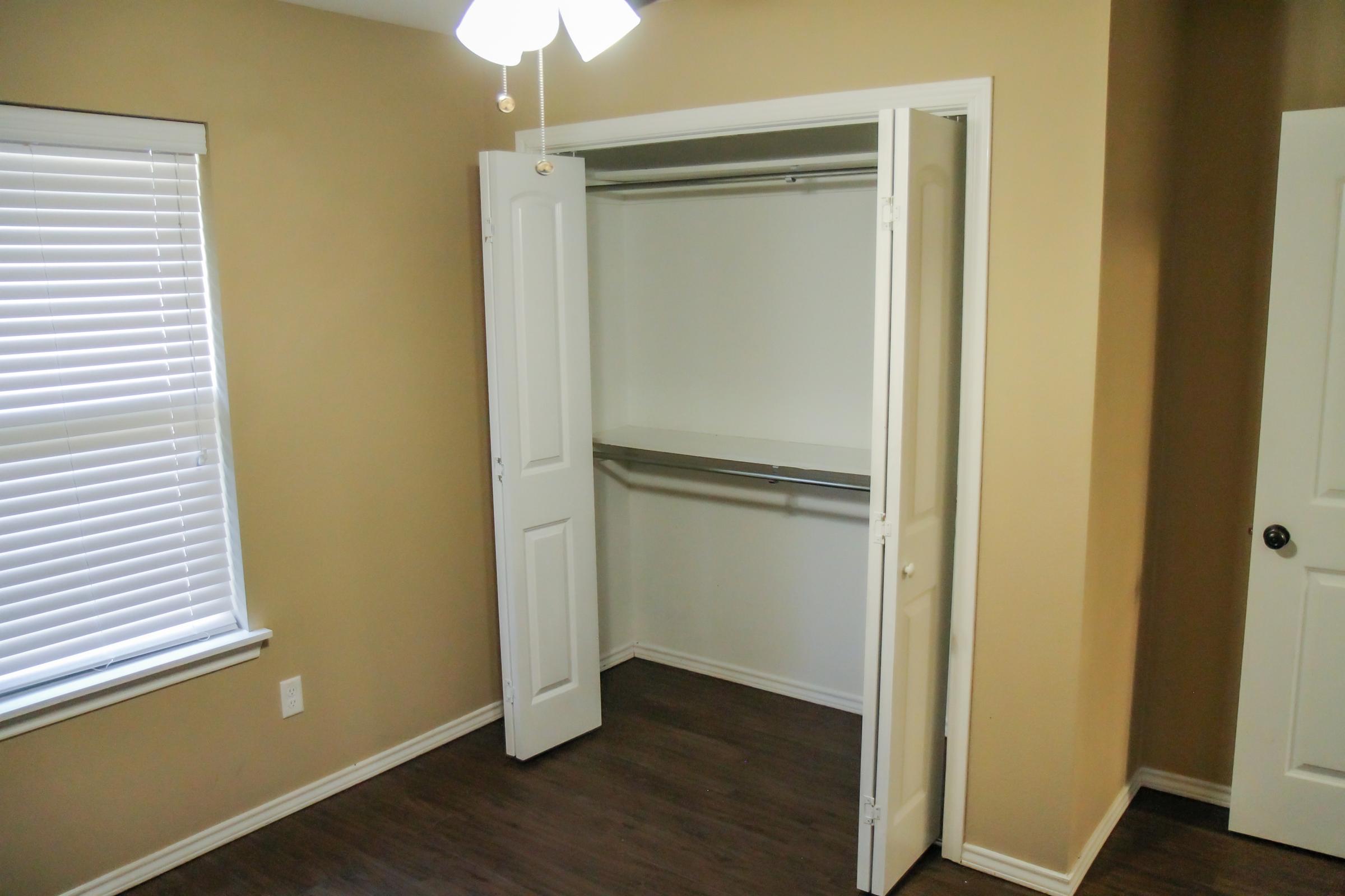 A spacious, empty closet with double doors, featuring a hanging rod and a plain white interior. The room has light brown walls and a ceiling fan, with a window to the left allowing natural light to enter. The floor is dark wood, creating a contrast with the walls.