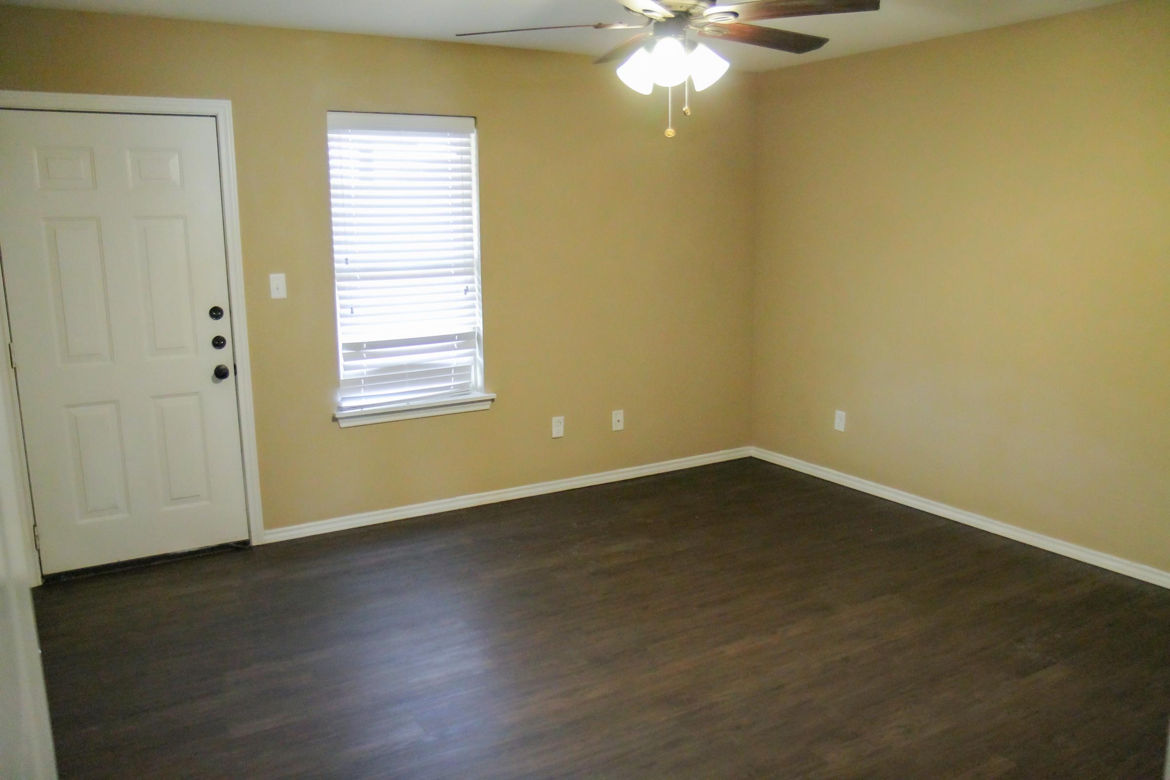 A vacant room with light brown walls and dark wood-like flooring. There is a white door on the left, a window with blinds on the right, and a ceiling fan in the center. The space is well-lit and has minimal furnishings, creating an open and airy atmosphere.