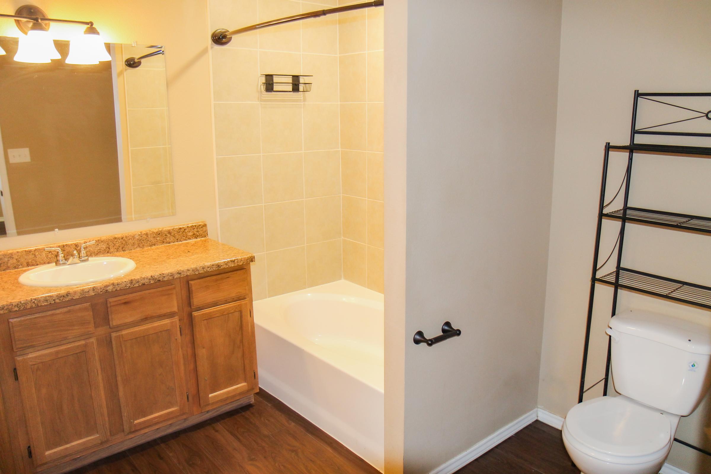 A clean bathroom featuring a bathtub with beige tiles, a wooden vanity with a sink, and a large mirror above it. To the right, there is a toilet next to a simple metal shelving unit. The walls are painted a light color, and there are two light fixtures above the mirror.