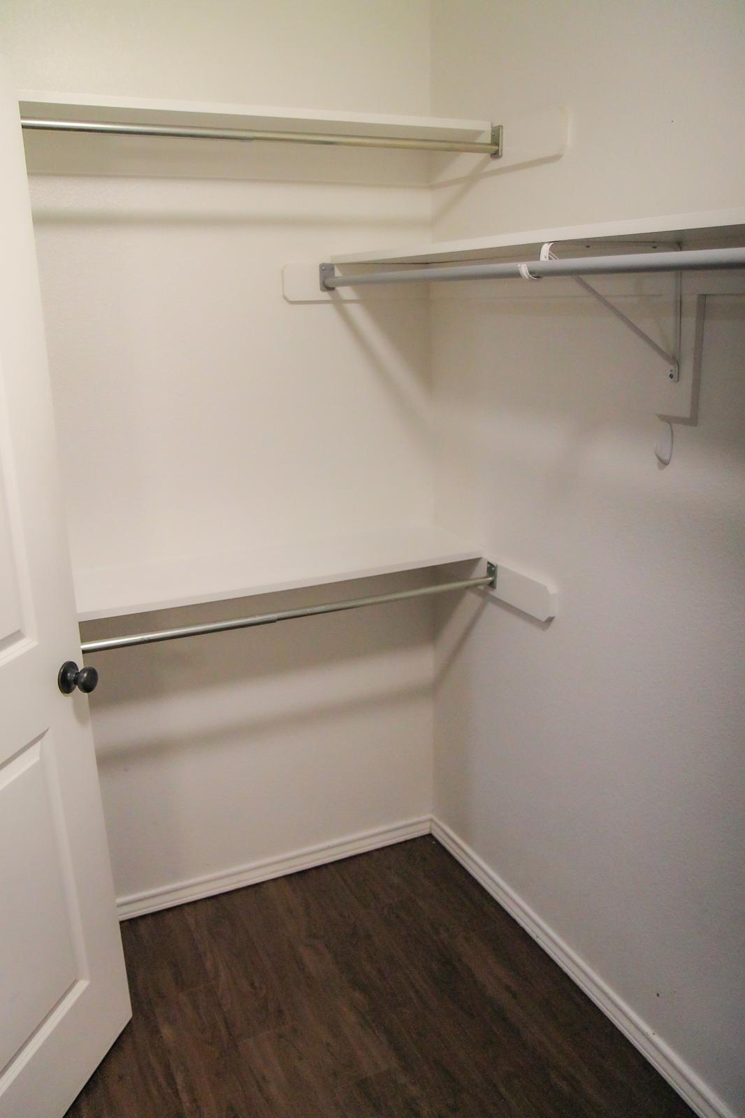 A minimal empty closet with white walls and two metal shelves mounted on the sides. There is a wooden floor and an open door on the left side, creating a clean and spacious appearance.