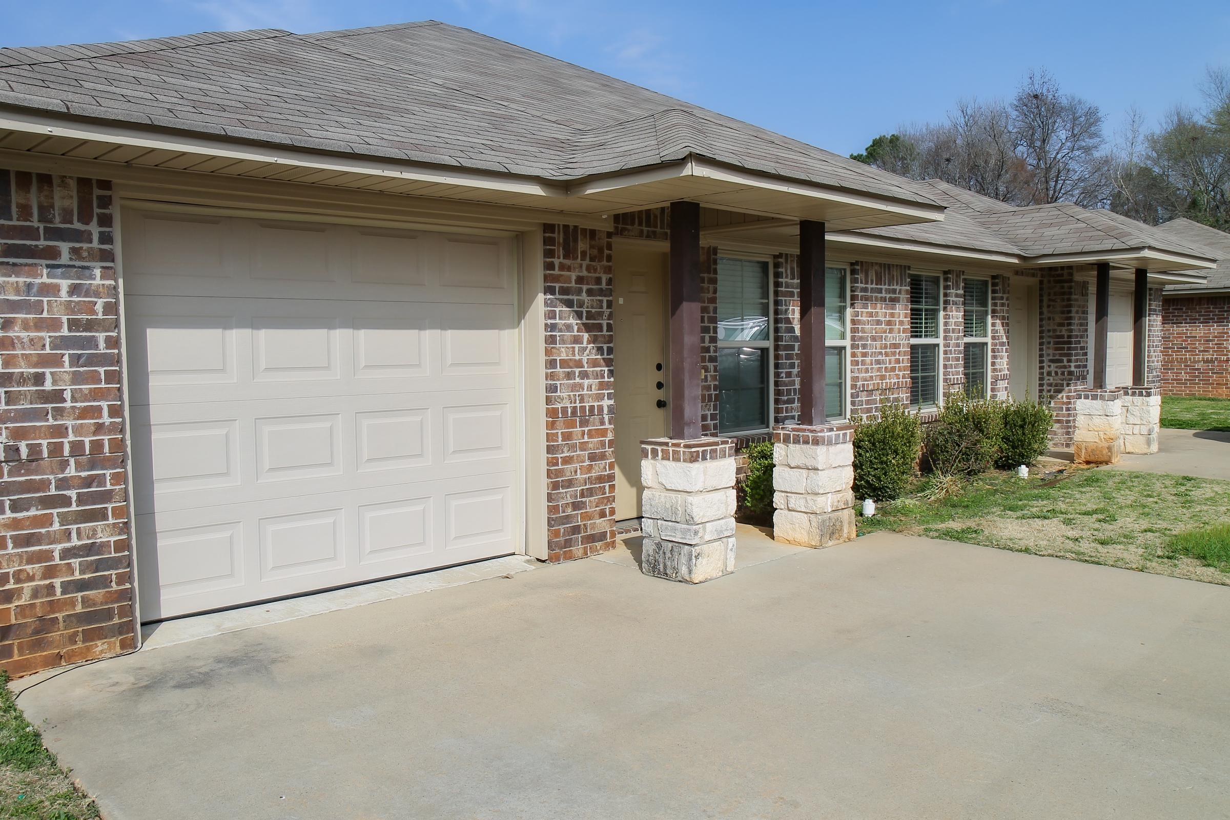 A residential house with a brick exterior and a beige garage door. The house has a concrete driveway and a small lawn with shrubs in front. It features a single-story design with multiple windows and a simple roofline, set against a clear blue sky.