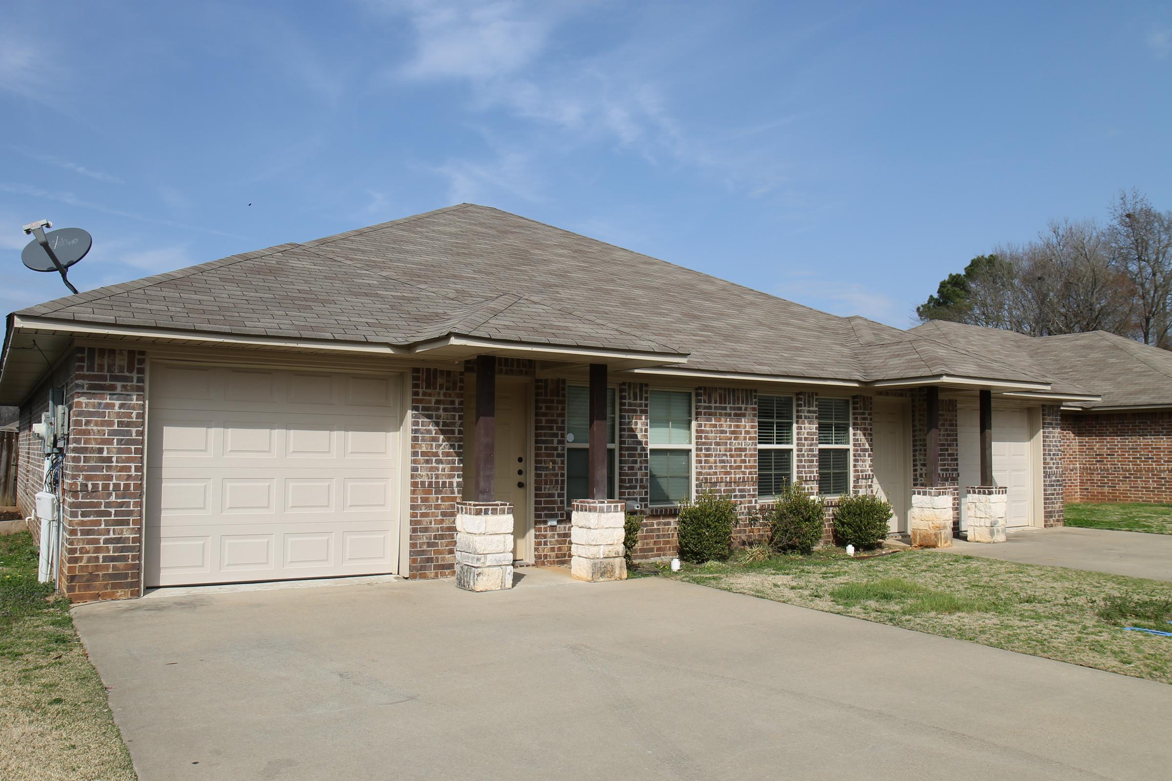 A single-story brick house with a garage and a paved driveway, featuring a front porch and several windows. The yard is neatly maintained with small bushes and grass. In the background, there are trees and a clear blue sky.