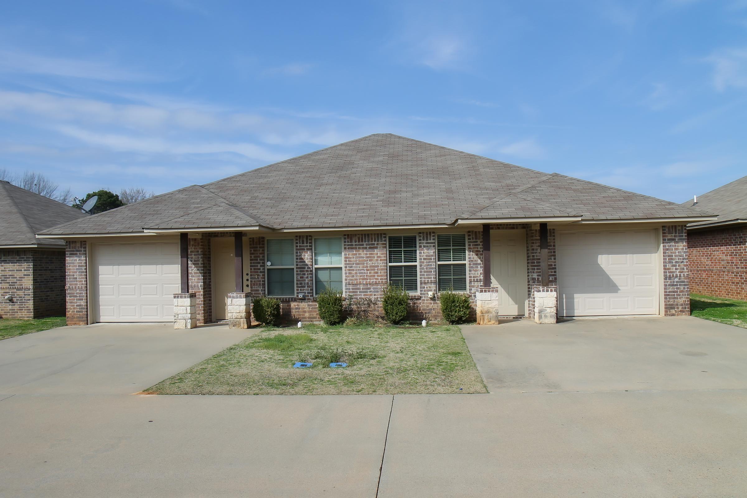 A view of a duplex with two separate entrances, featuring brick exteriors and garages. The building is set against a clear blue sky, and there is a small lawn with neatly trimmed bushes in front. The driveway is paved and leads up to the garages on either side.