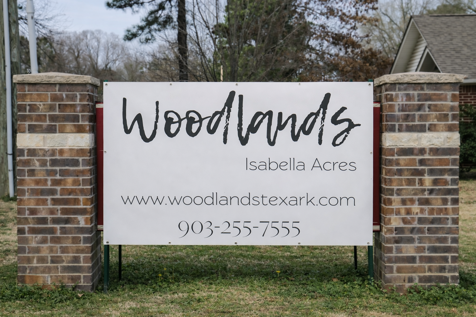 Sign displaying "Woodlands" and "Isabella Acres," with a website (www.woodlandstexark.com) and phone number (903-255-7555). The sign is mounted on a brick structure, set against a backdrop of trees and grassy area.