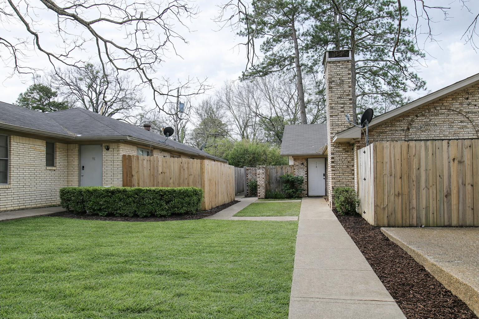 A view of a residential area featuring two single-story brick houses with a grassy lawn, well-kept shrubbery, and paved walkways. Between the houses, there's a wooden fence. The sky is partially cloudy, and trees are visible in the background.