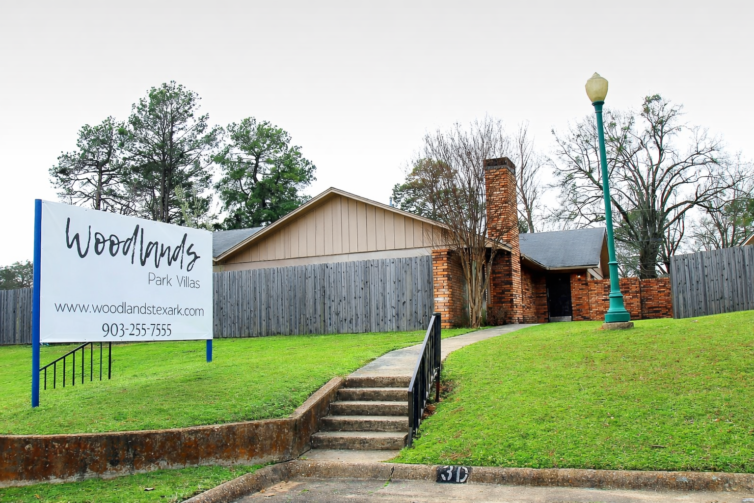 A brick building with a pitched roof is set back from a landscaped lawn. A sign reads "Woodlands Park Villas" with contact information. In front, a curved walkway leads to a low brick wall, and a lamppost stands nearby. Trees can be seen in the background, indicating a calm residential area.