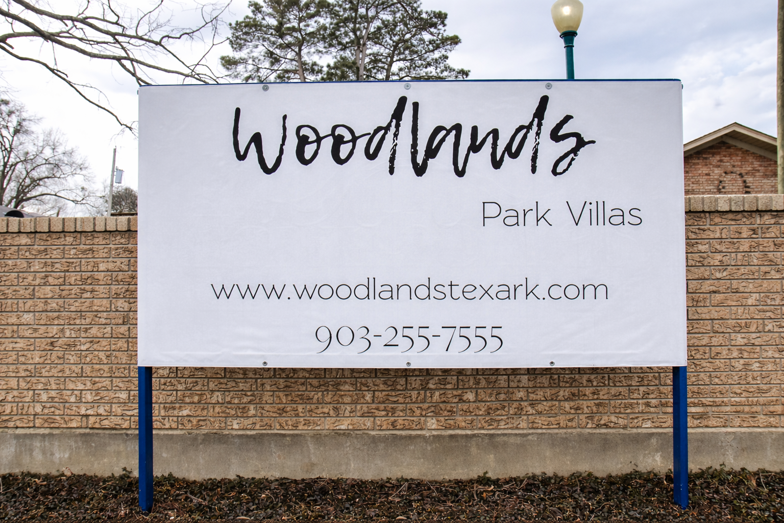 Sign for Woodlands Park Villas featuring the website URL www.woodlandstexark.com and a contact number, 903-255-7555. The sign is displayed on a brick wall with a natural setting in the background.