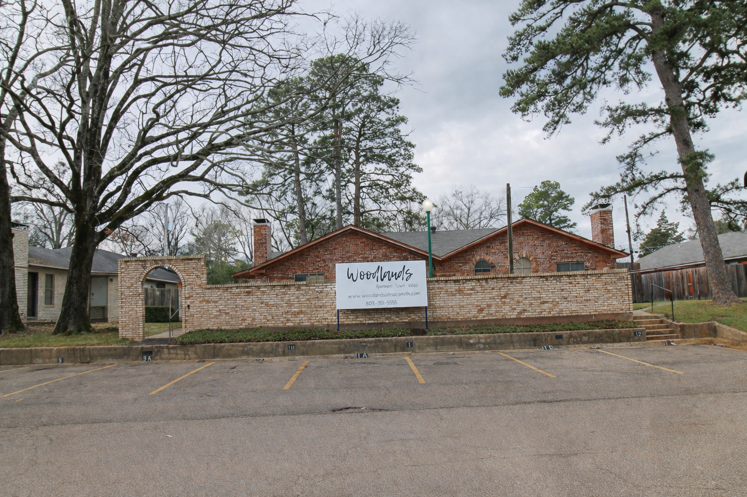 A brick building with a large sign that reads "Woodlands" is set in front of a tree-lined area. The building features two chimneys and is surrounded by a low wall. The sky is overcast, and there is a parking area in front with multiple numbered spaces.