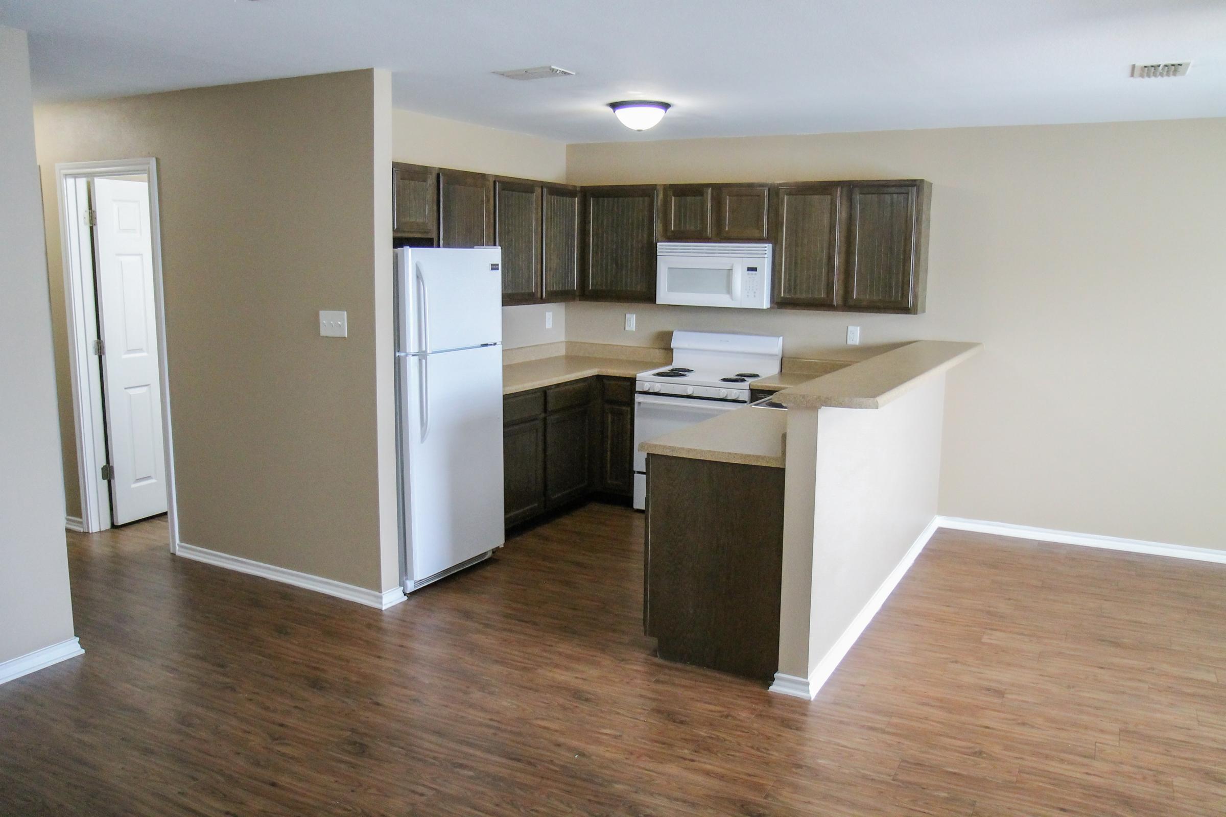 Modern kitchen and living area featuring brown wood cabinetry, white appliances including a refrigerator and stove, light beige walls, and laminate flooring. A doorway leads to another room, enhancing the open layout.