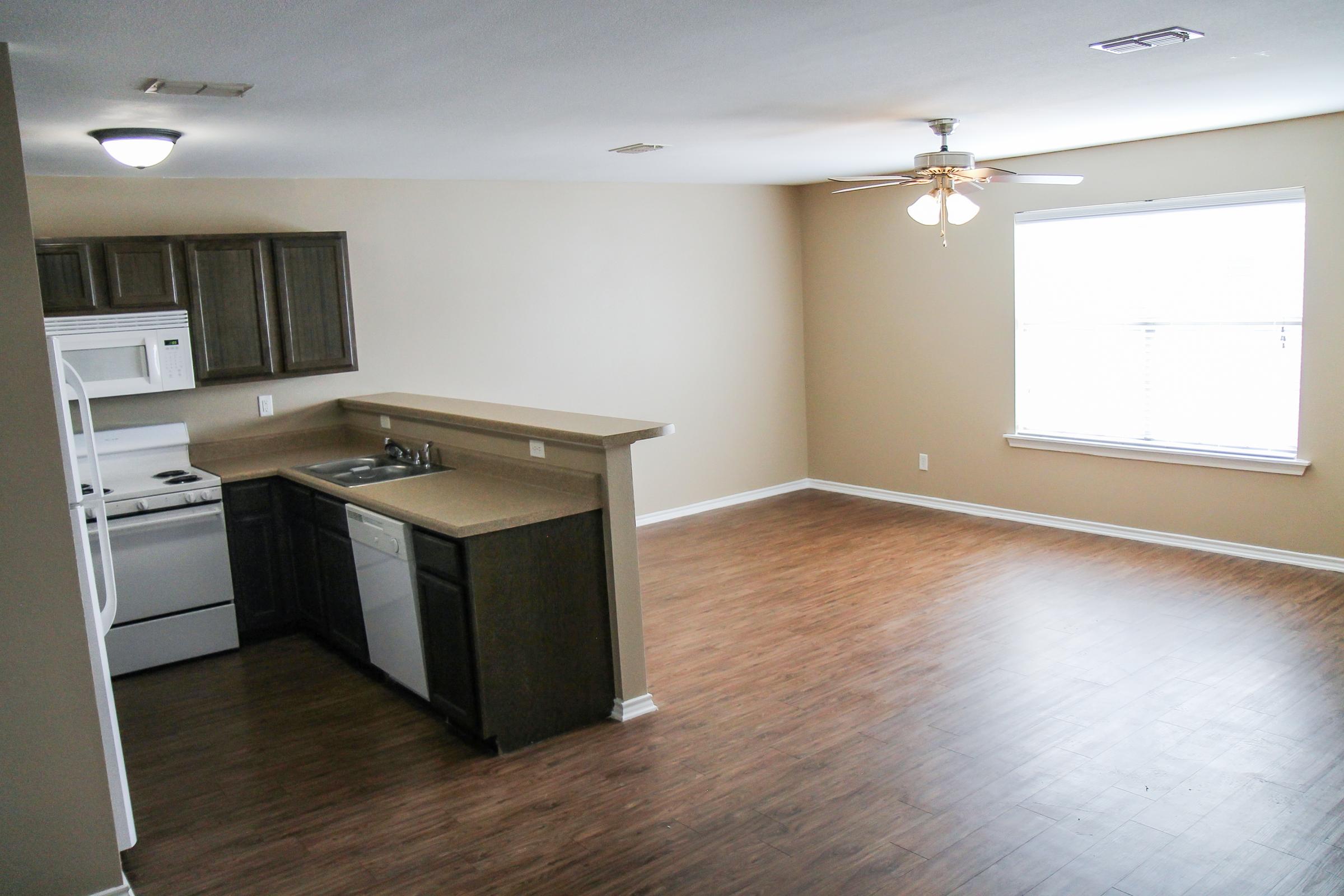 An empty interior of an apartment featuring a kitchen area with a white refrigerator, stove, and dishwasher. The open living space has light-colored walls and laminate flooring, with a ceiling fan and a large window providing natural light.