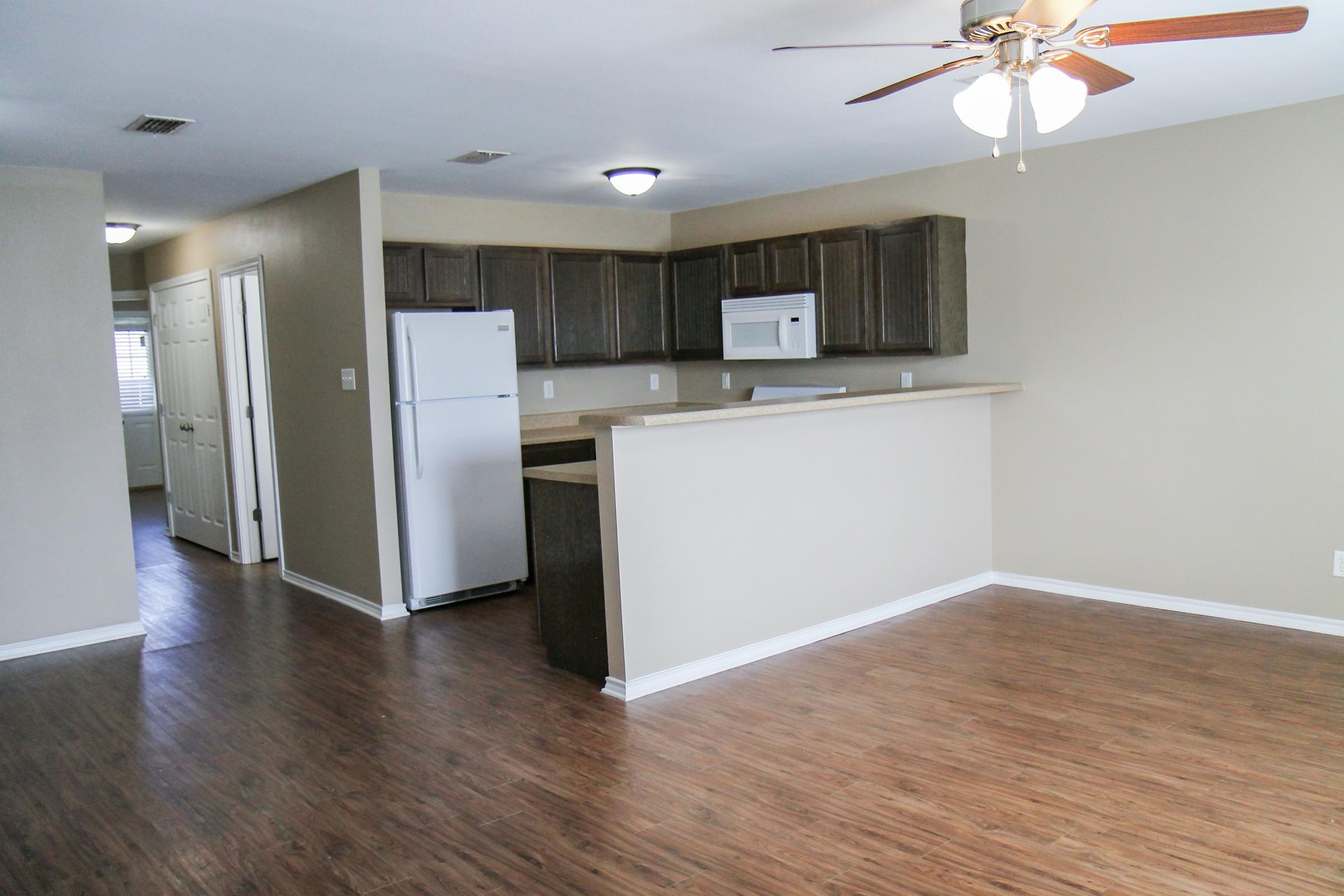 A spacious, unfurnished living area with a wooden floor, featuring a ceiling fan, an open kitchen with dark wooden cabinets, and a white refrigerator. The room is well-lit, has a neutral-colored wall, and connects to a hallway leading to other rooms.