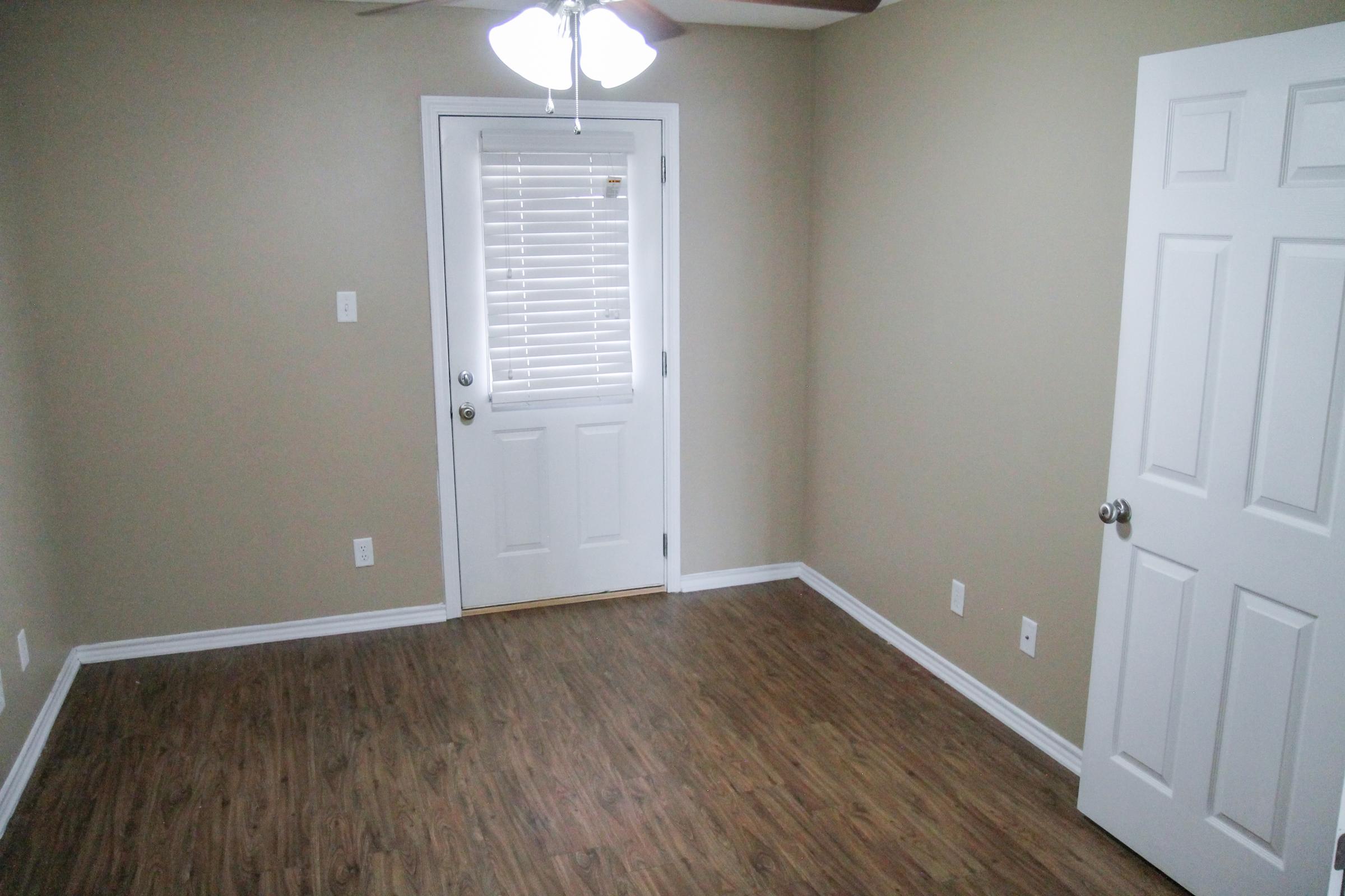 A small, empty room with light brown walls and laminate flooring. There's a ceiling fan and a white door leading outside, with a window beside it covered by blinds. Another white door is visible on the right side of the image, adding to the room's minimalistic design.