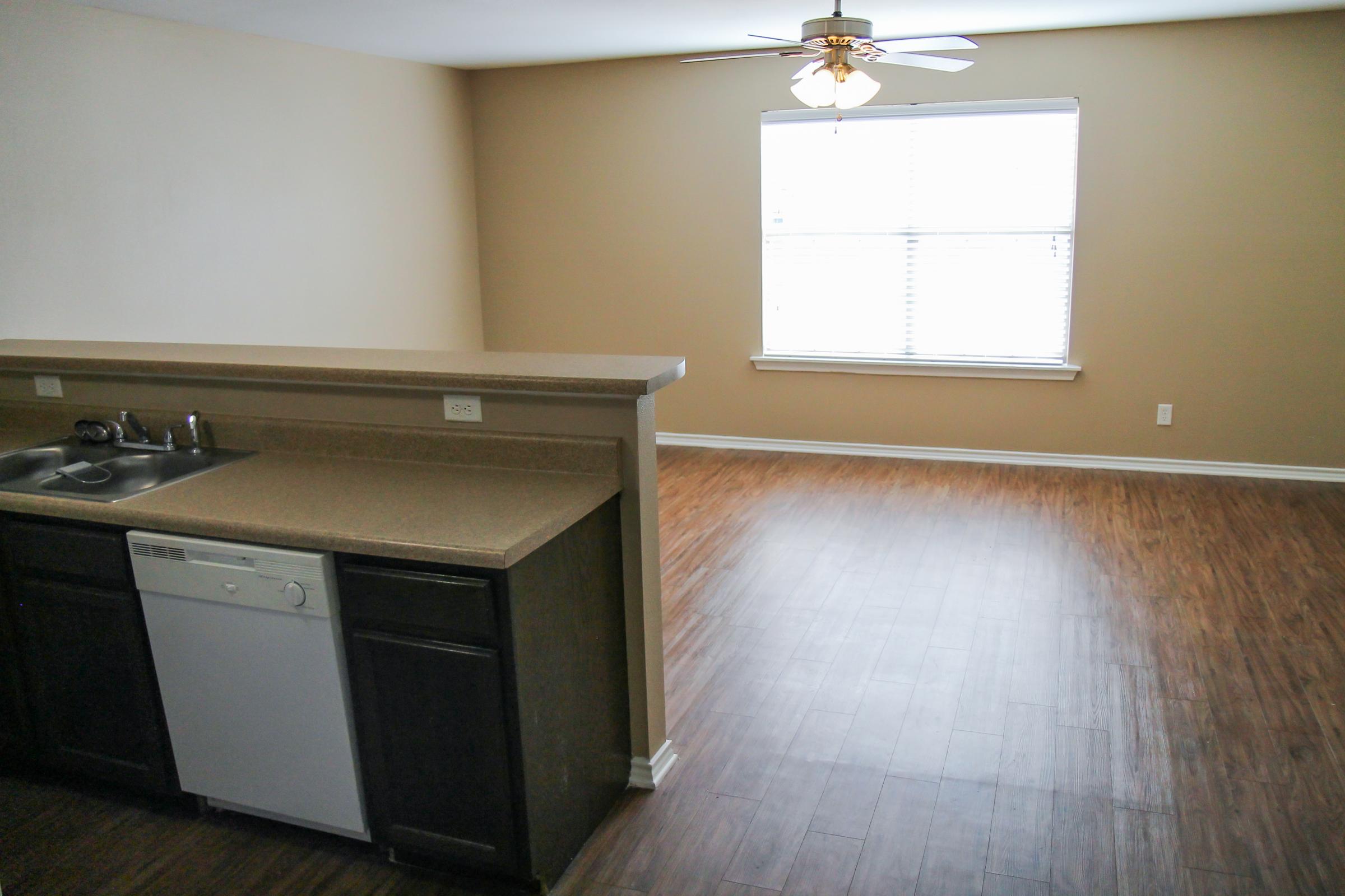 A spacious, well-lit living area with light brown walls and hardwood flooring. The kitchen area features a countertop with a sink and a dishwasher, while a large window allows natural light into the room. A ceiling fan hangs above, adding to the open layout.