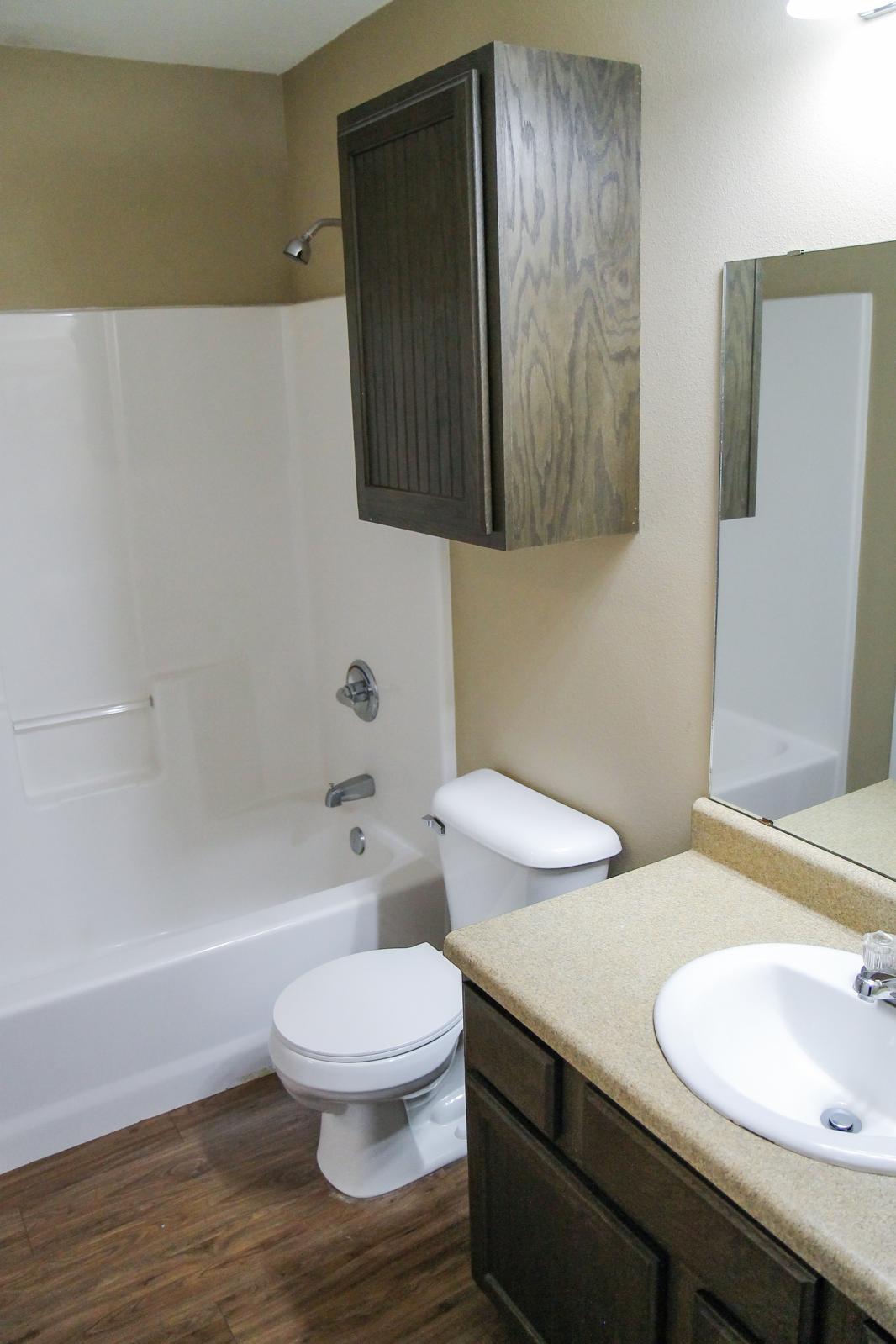 A modern bathroom featuring a white bathtub and shower combination, a white toilet, and a countertop with a sink. The walls are painted a neutral color, and there is a dark wooden cabinet above the toilet. A mirror is mounted above the sink, and the flooring is a wood-like laminate.