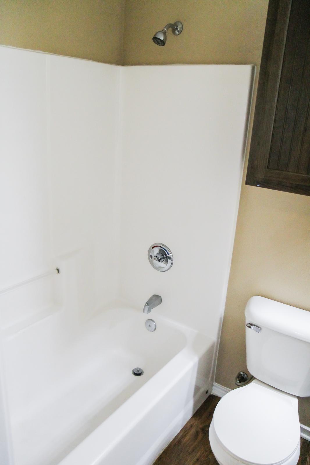 A clean, modern bathroom featuring a white shower/tub combo on the left, a chrome faucet and showerhead, and a dark wood cabinet above. To the right, there's a white toilet next to a light, neutral wall. The floor is covered in a wooden finish, creating a warm atmosphere.