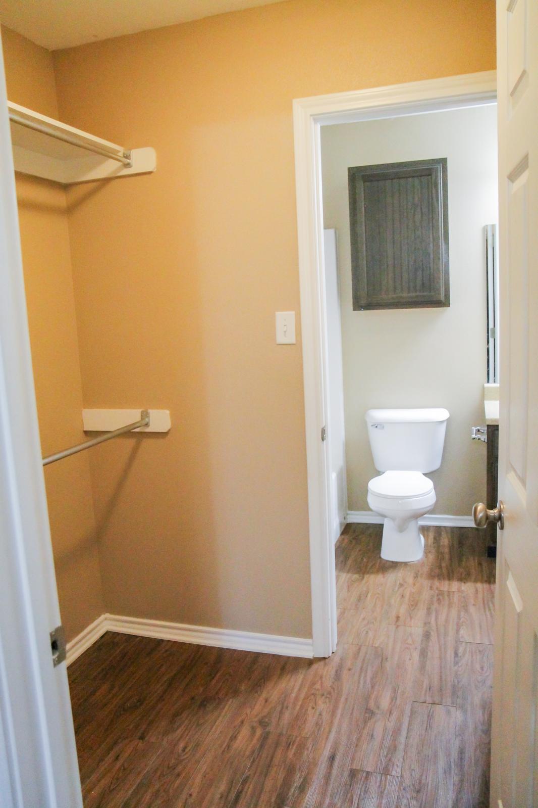 A small bathroom with a toilet visible through an open door. Adjacent to it is a closet area with a hanging rod and light brown walls. The floor features wood-like tiles, and there is a utility storage cabinet mounted on the wall in the bathroom. The overall space is neatly organized and well-lit.