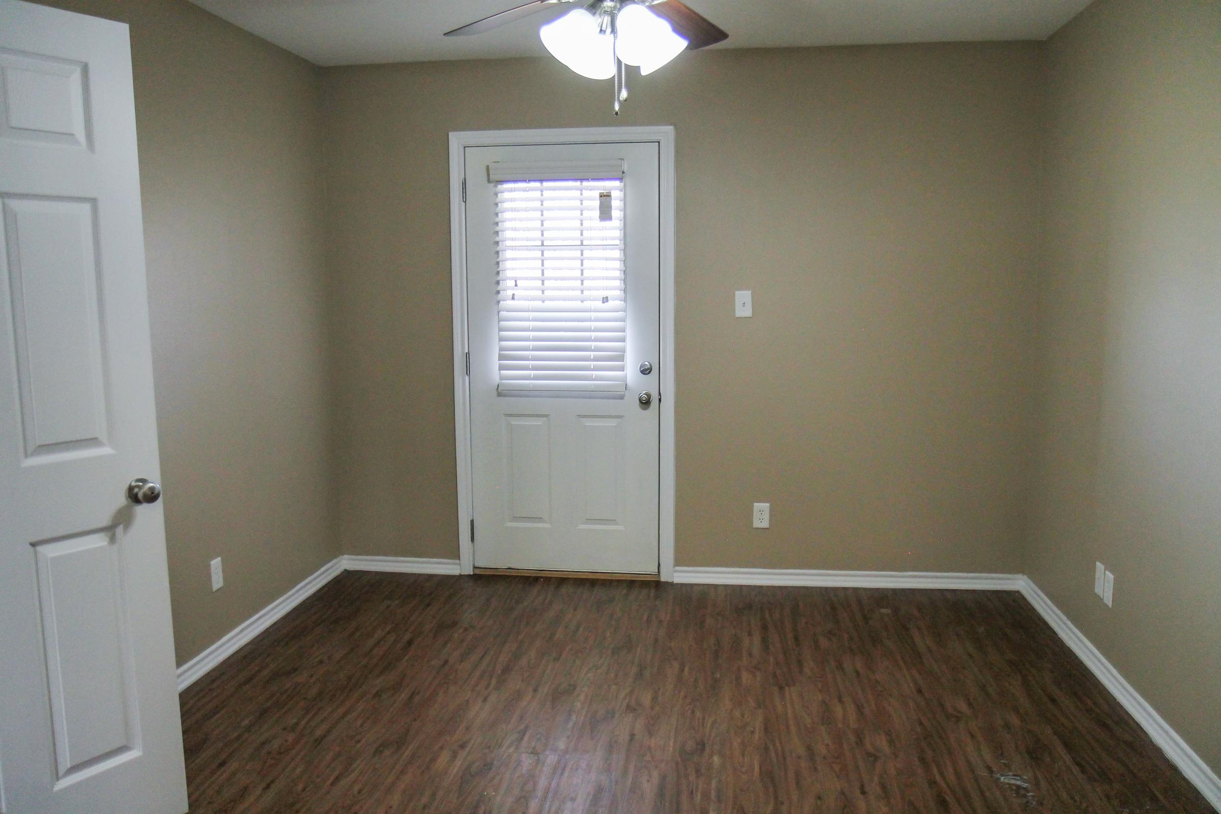 A small, empty room with light brown walls and wooden flooring. There's a closed white door on the left and a white door leading outside on the right, complete with a window covered by blinds. A ceiling fan hangs above, adding a touch of brightness to the space.