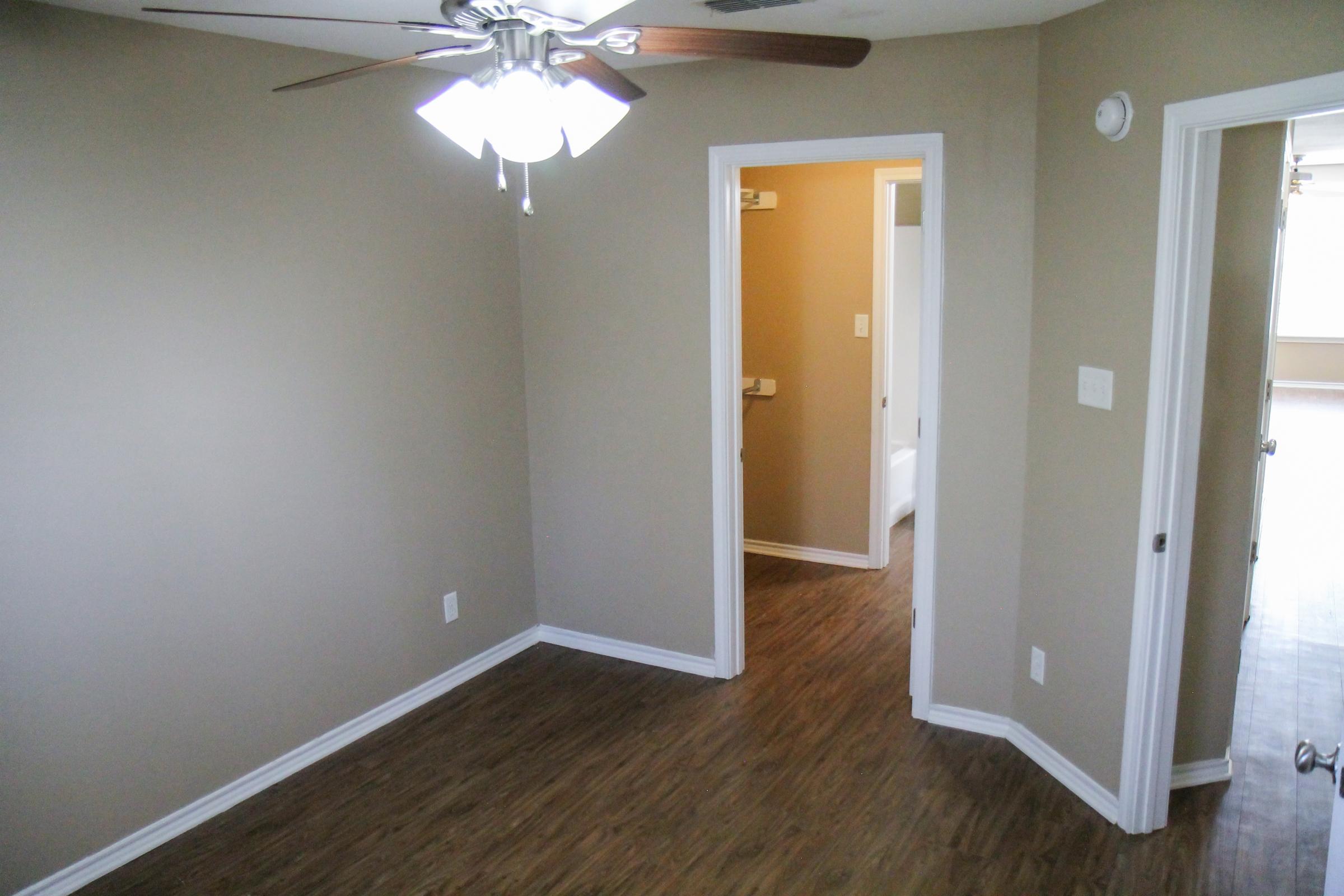 A simple, well-lit room featuring a ceiling fan with light fixtures. The walls are painted a light brown, and there are two doorways: one leading to a bathroom and another out of the room. The floor is covered with a wooden laminate, creating a cozy atmosphere.
