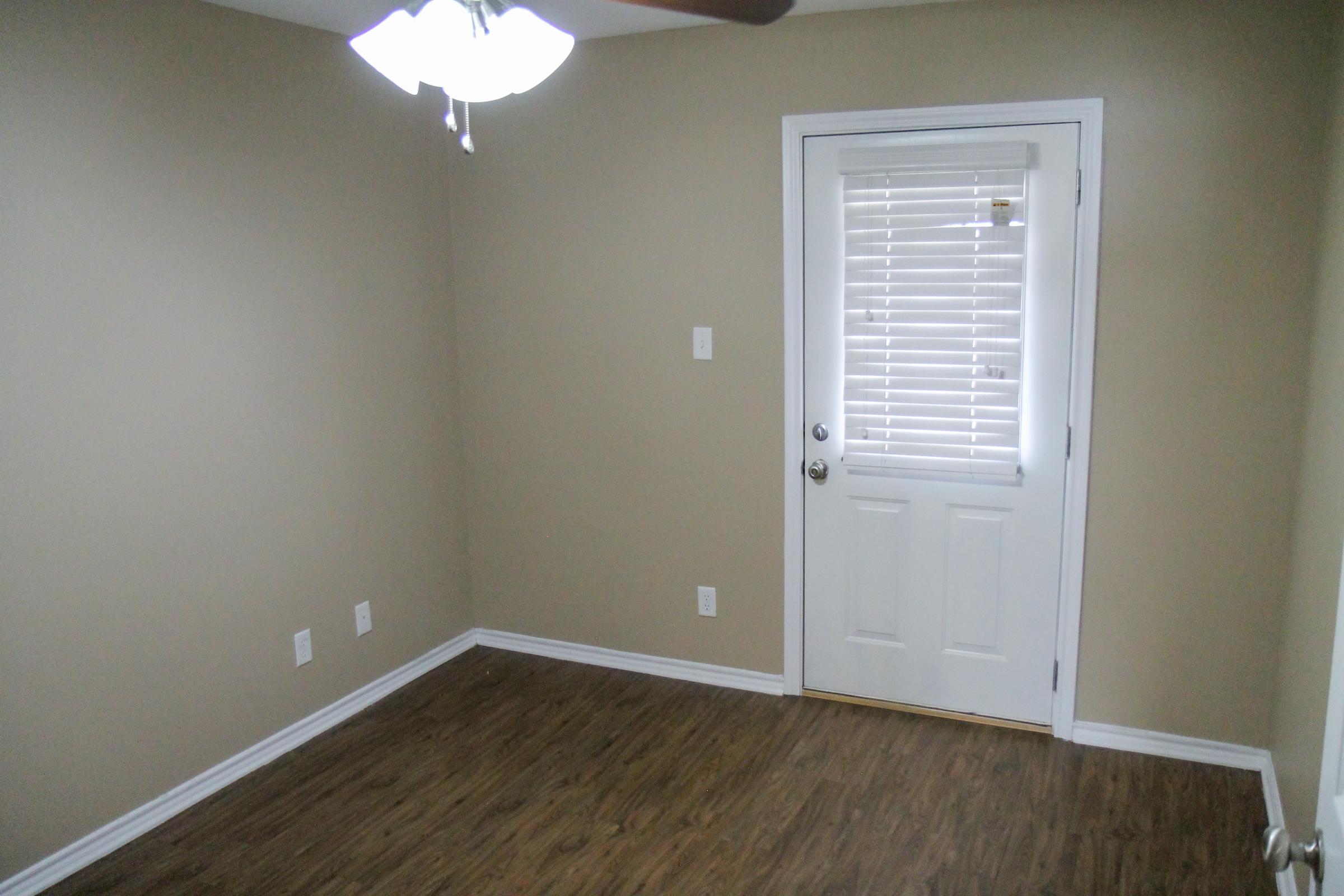 A small, empty room with light brown walls and a light wood floor. A ceiling fan is visible, and there's a door with a window that has blinds. The room has minimal furnishings and appears well-lit, conveying a clean and simple aesthetic.