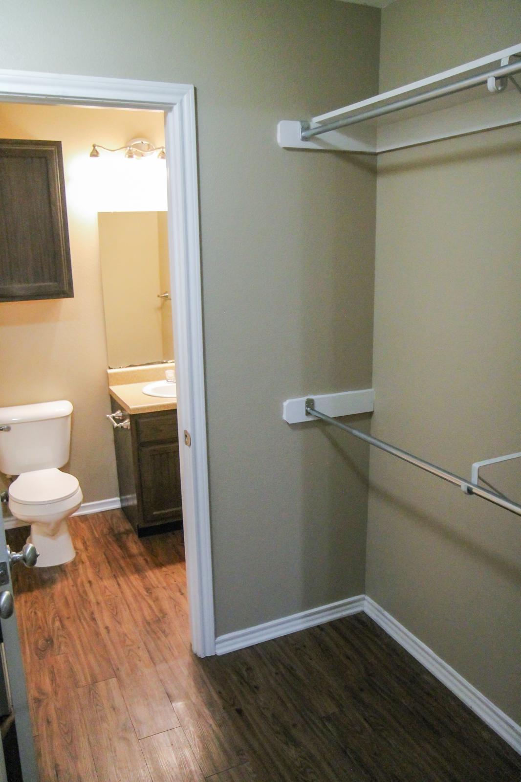 A view of a bathroom and adjoining closet area. The bathroom features a toilet and a vanity with a sink, while the closet has a hanging rod and shelves. The walls are painted in a neutral color, and the floor is wood-like laminate.