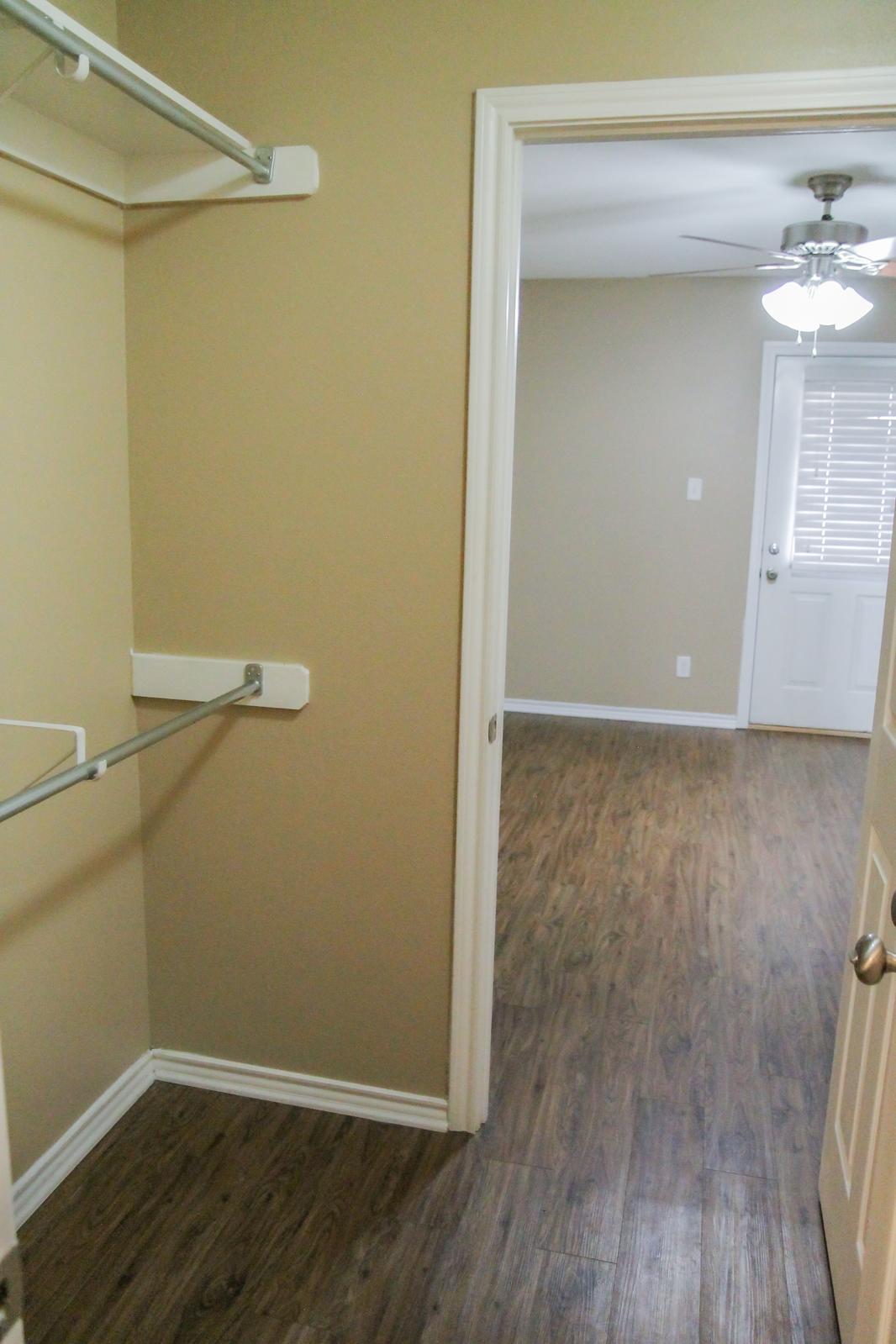 A view of a small room showcasing a closet with a single hanging rod and a doorway leading into a larger room. The larger room features a ceiling fan and a door with a window. The walls are painted a neutral beige, and the flooring is a dark wood laminate.