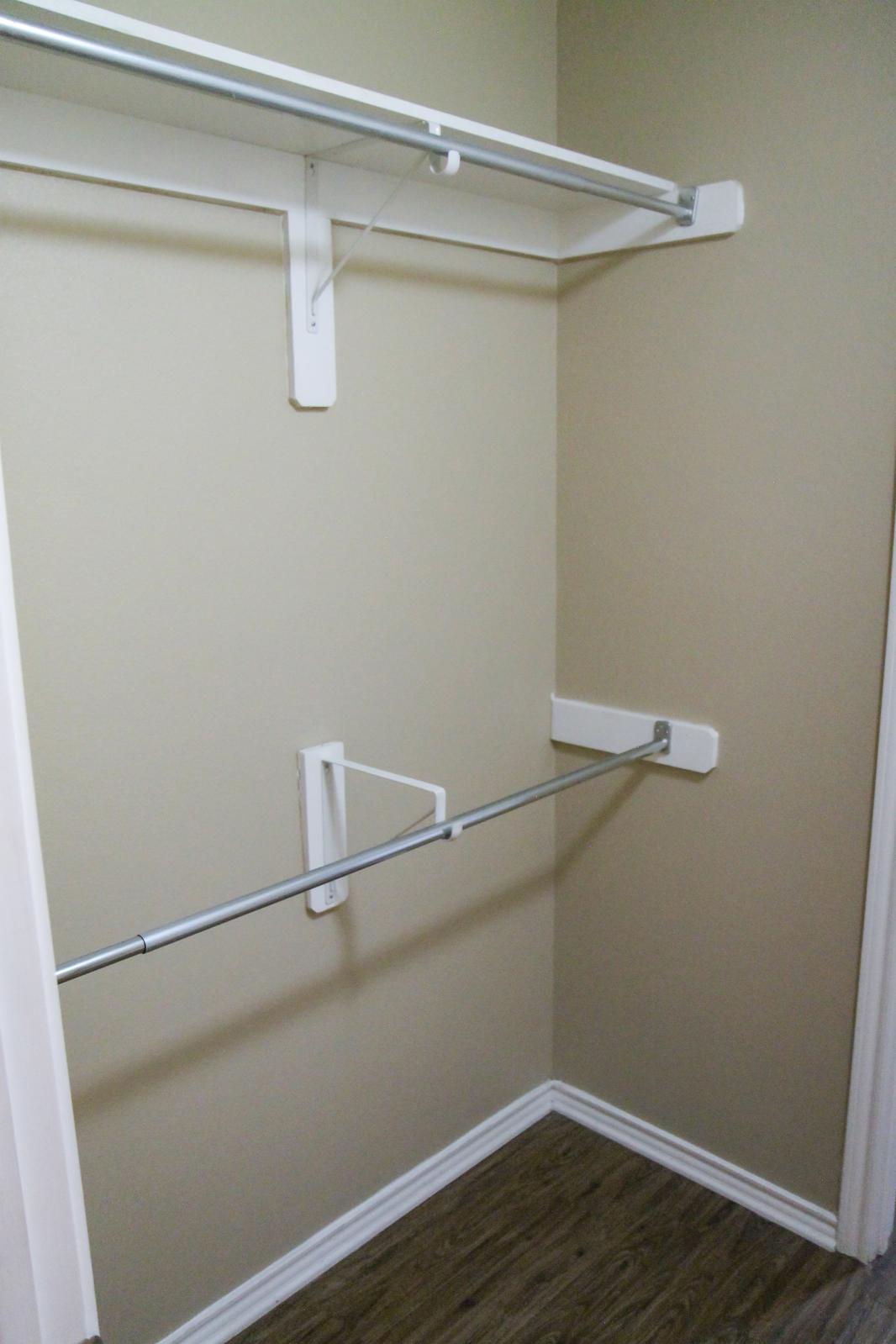 Empty closet with two horizontal shelves mounted on the walls and a single clothes rod in the middle. The walls are painted a light color, and the floor features laminate or hardwood flooring. The overall space is uncluttered and ready for storage or organization.