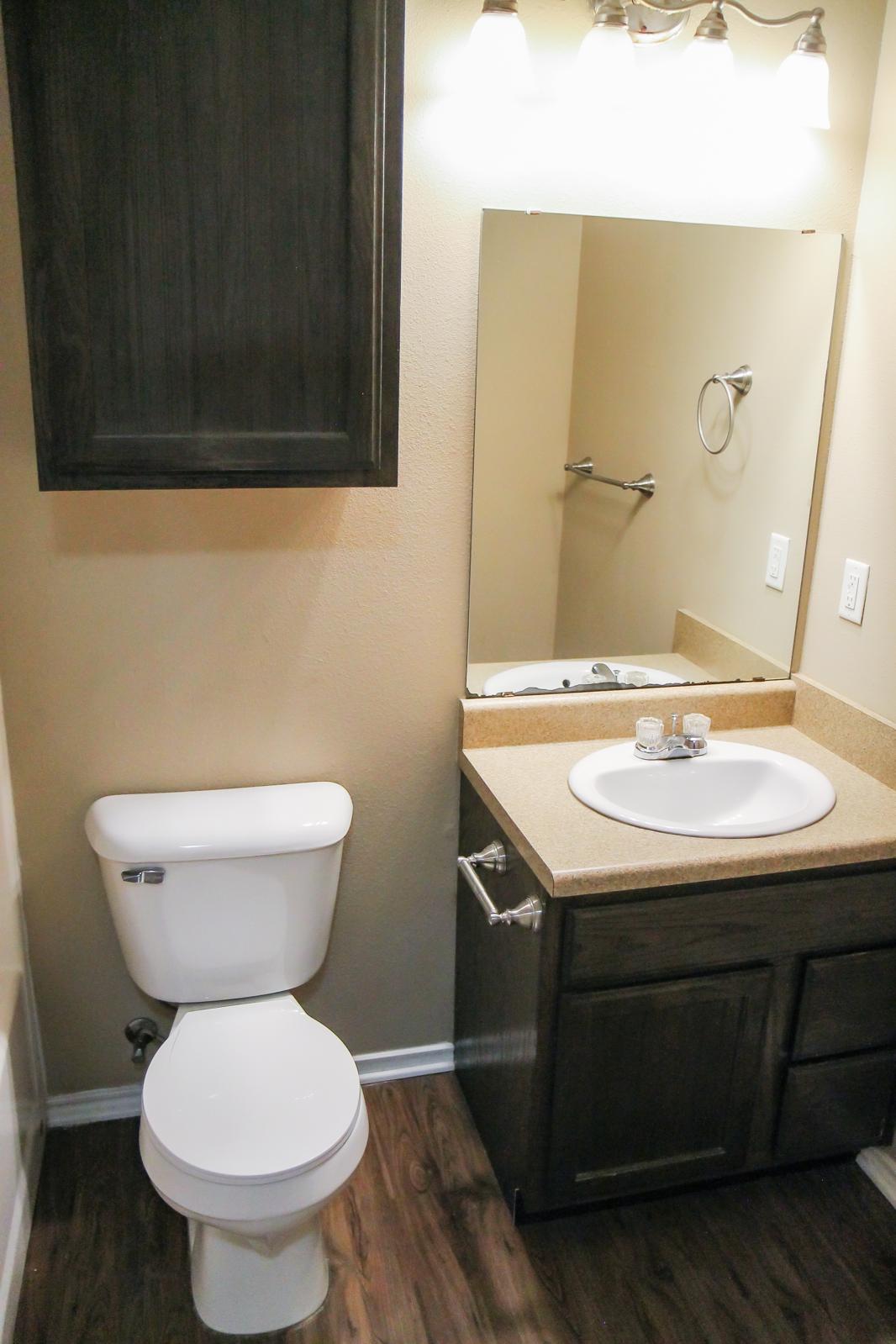 A small bathroom featuring a toilet, a sink with a countertop, a mirror above the sink, and a wall-mounted towel holder. The walls are painted a soft beige color, and there is a dark wooden cabinet above the toilet. The flooring is hardwood.