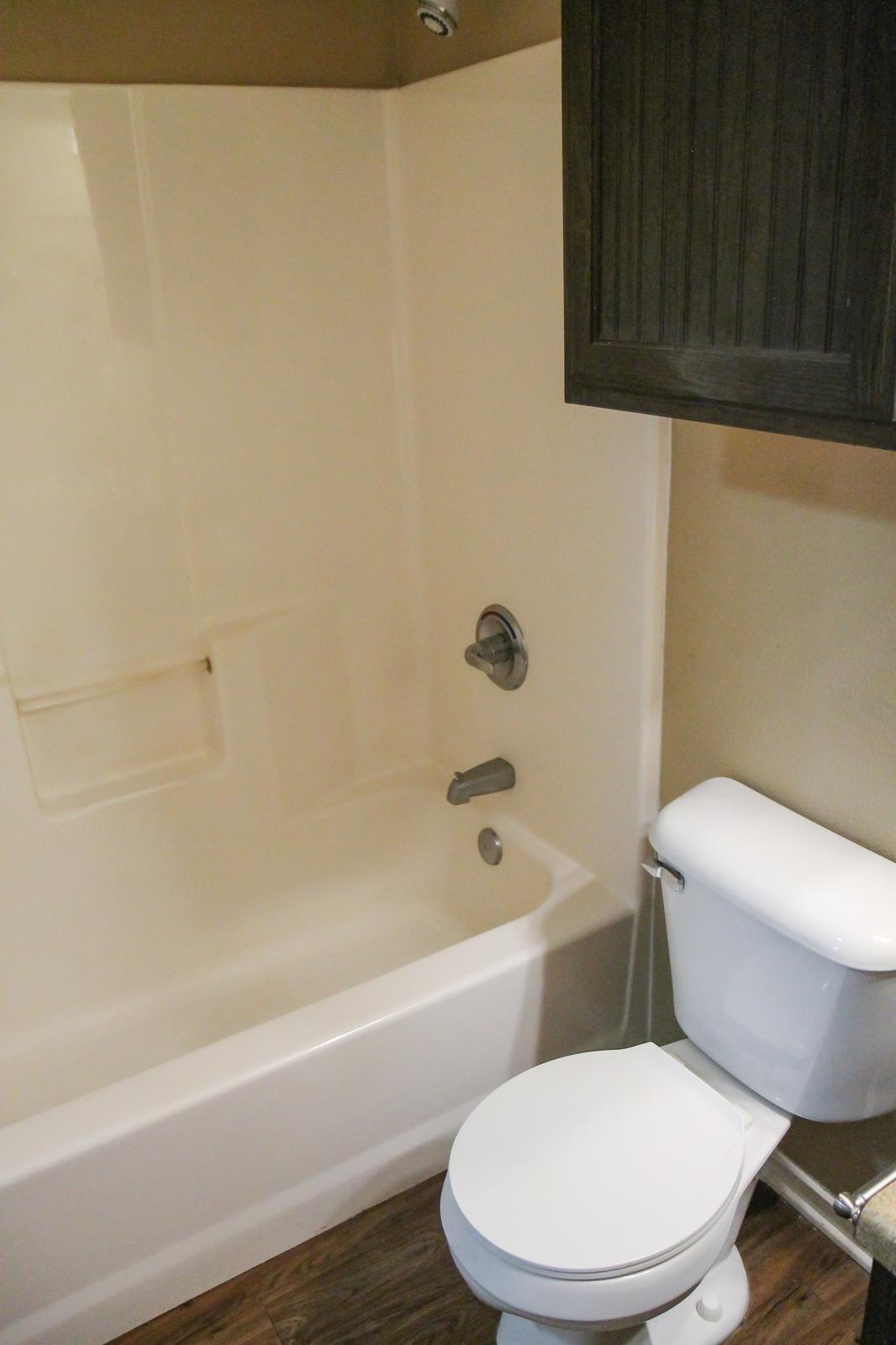 A bathroom featuring a bathtub with a shower fixture and a white toilet. The walls are a light neutral color, and there is a dark wood cabinet above the toilet. The floor is a dark wood laminate.