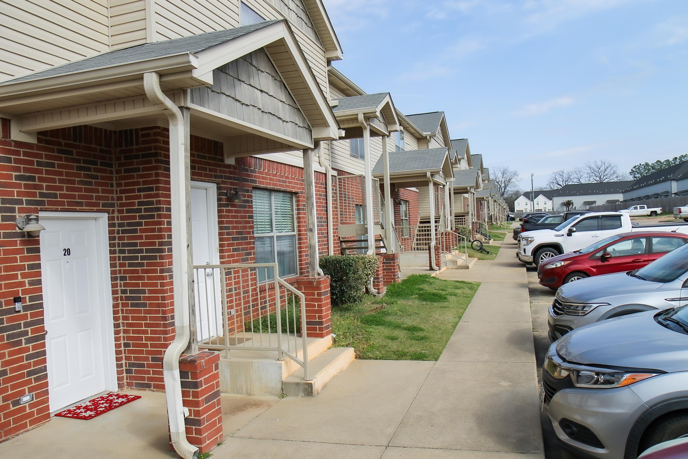 A row of multi-unit residential buildings featuring brick facades and small front porches, with neatly trimmed bushes. The sidewalk is flanked by parked cars, including several sedans in various colors, on a clear day with blue skies.
