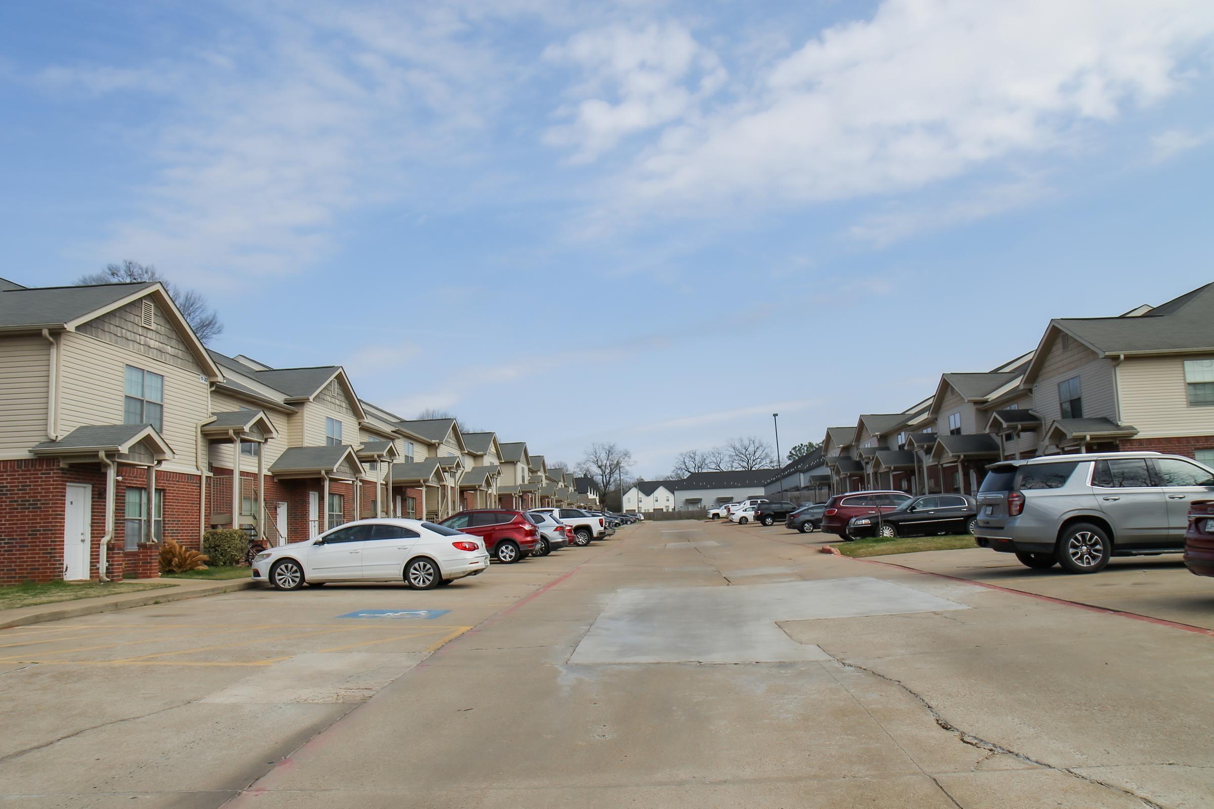 A residential street lined with multi-family homes, featuring parking spaces filled with various cars. The sky is bright with scattered clouds, and some greenery is visible near the buildings. The scene conveys a calm, suburban atmosphere.